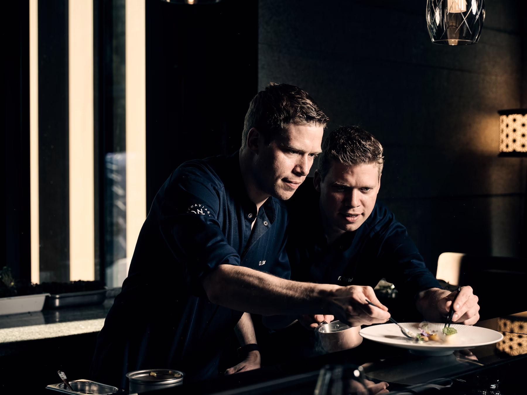 Zwei Köche arrangieren sorgfältig feine Speisen auf einem Teller in einer dunklen, stilvollen Restaurantküche.