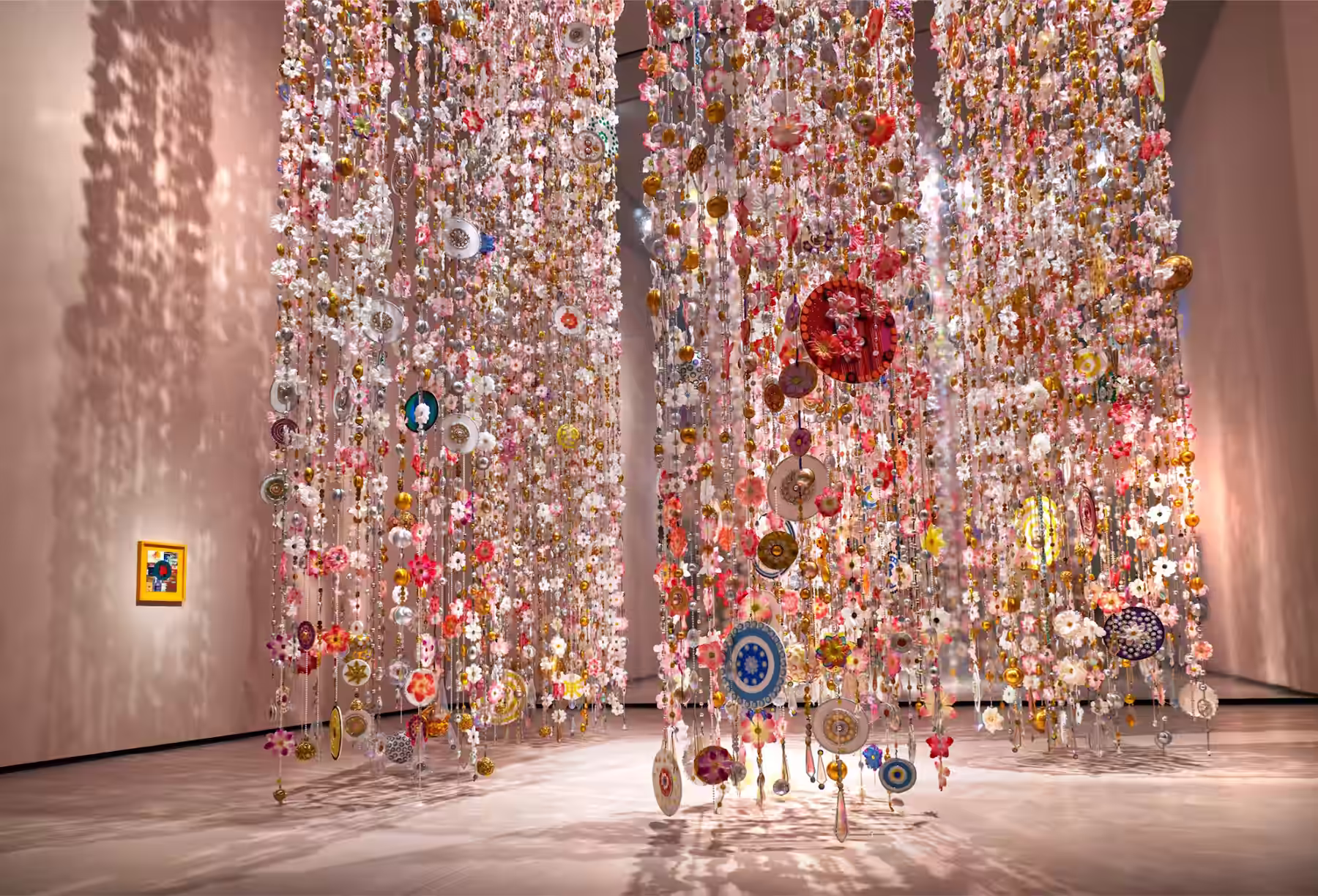 Bunte hängende Kunstinstallation aus zahlreichen feinen Perlen, Blumen und kleinen Scheiben, die Schatten an die Wände eines geräumigen Raums werfen.