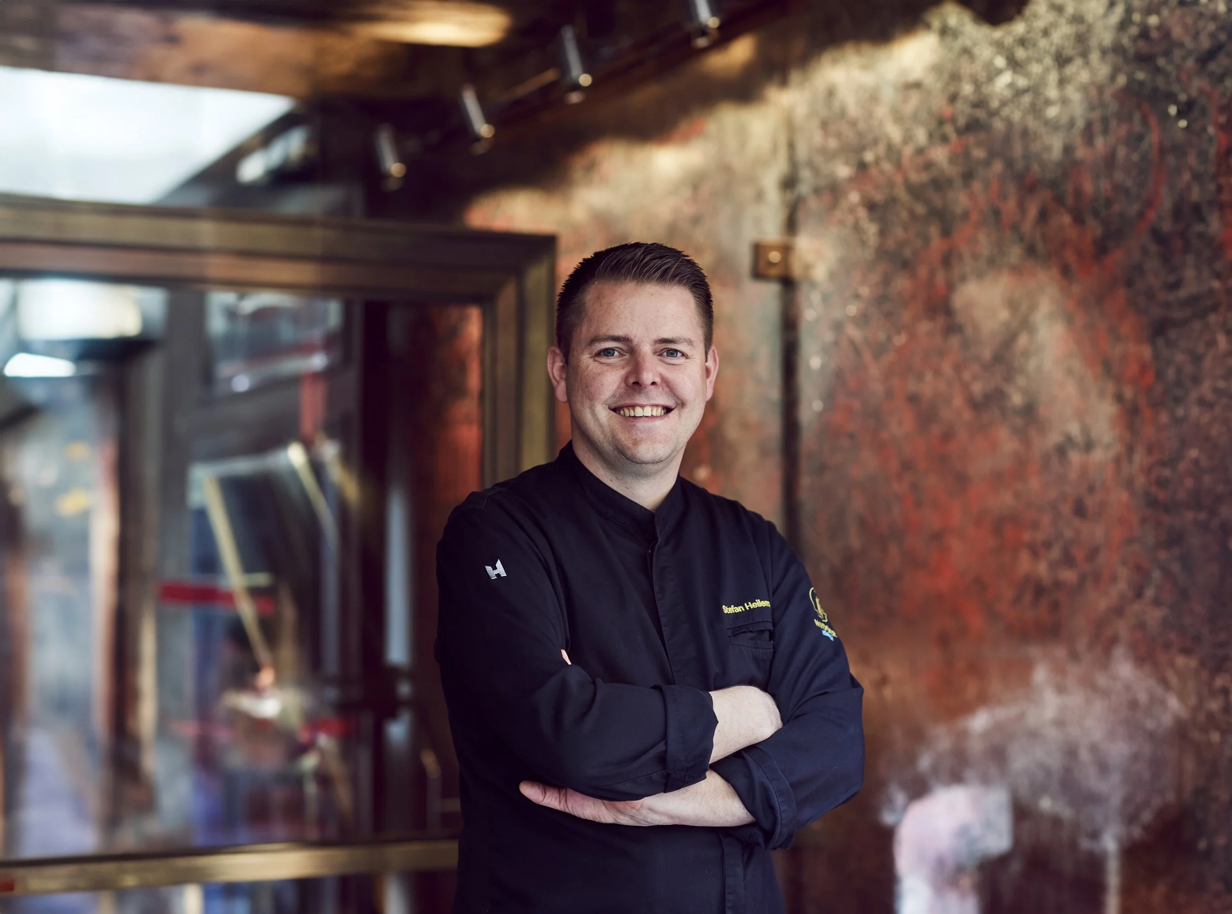 Lächelnder Mann in schwarzer Kochjacke mit verschränkten Armen vor einer rustikalen Wand.