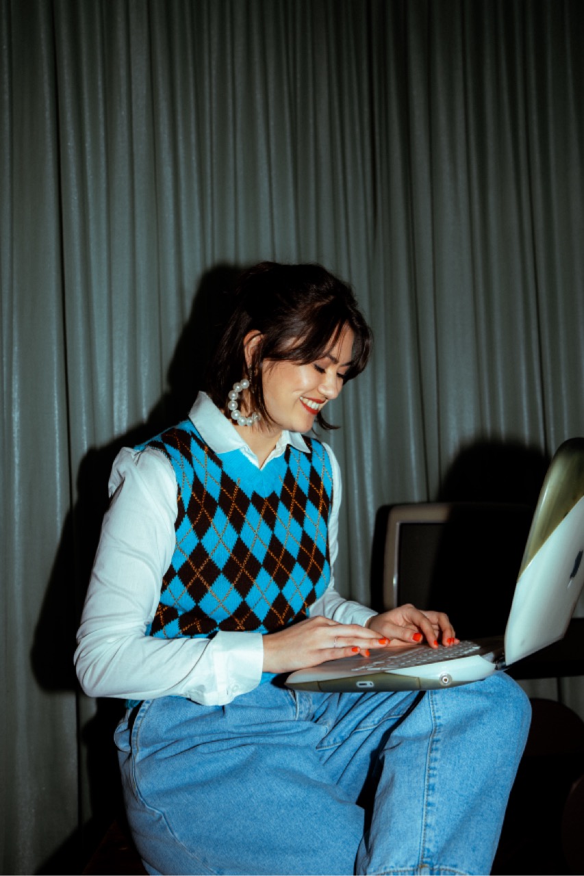 Vanessa im Retro-Look mit Argyle-Pullunder, tippend auf einem alten durchsichtigen Apple-Laptop, im Hintergrund ein Röhrenmonitor.