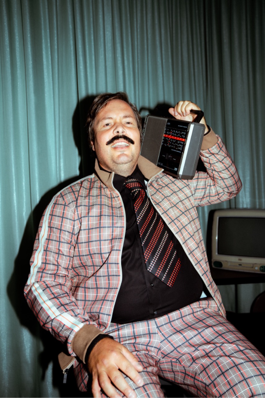 Benedikt im karierten Trainingsanzug mit Krawatte, hält ein altes Transistorradio ans Ohr.