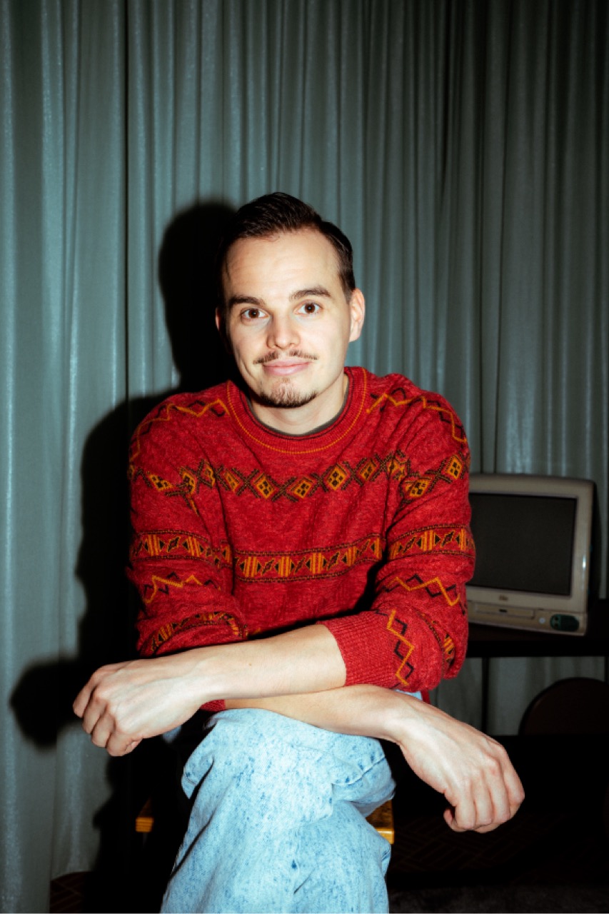 Dominic im rot gemusterten Pullover, sitzend mit verschränkten Armen.