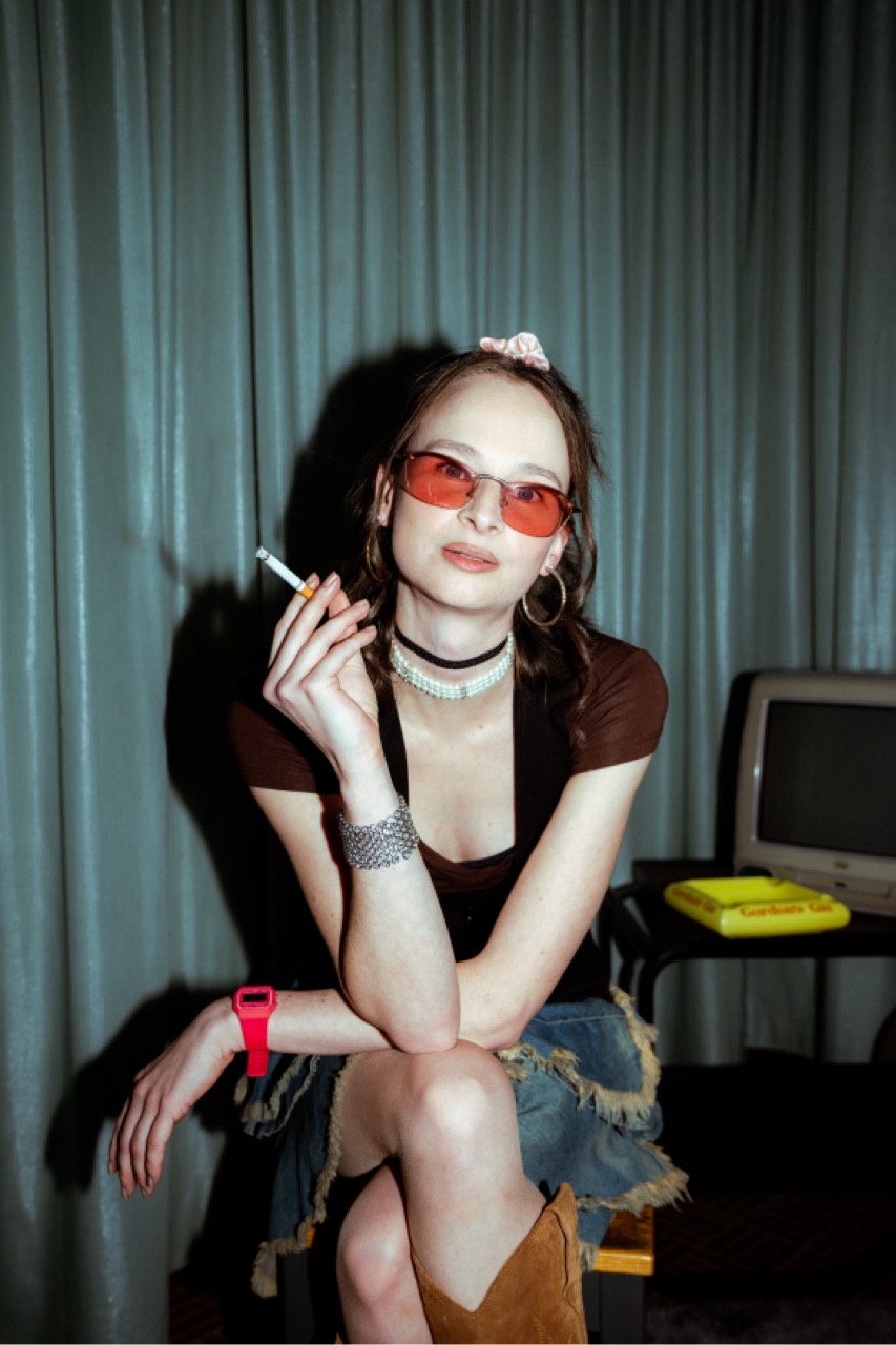 Elena mit getönter Sonnenbrille und Zigarette in der Hand, im ausgefransten Jeansrock, im Hintergrund ein alter Fernseher.