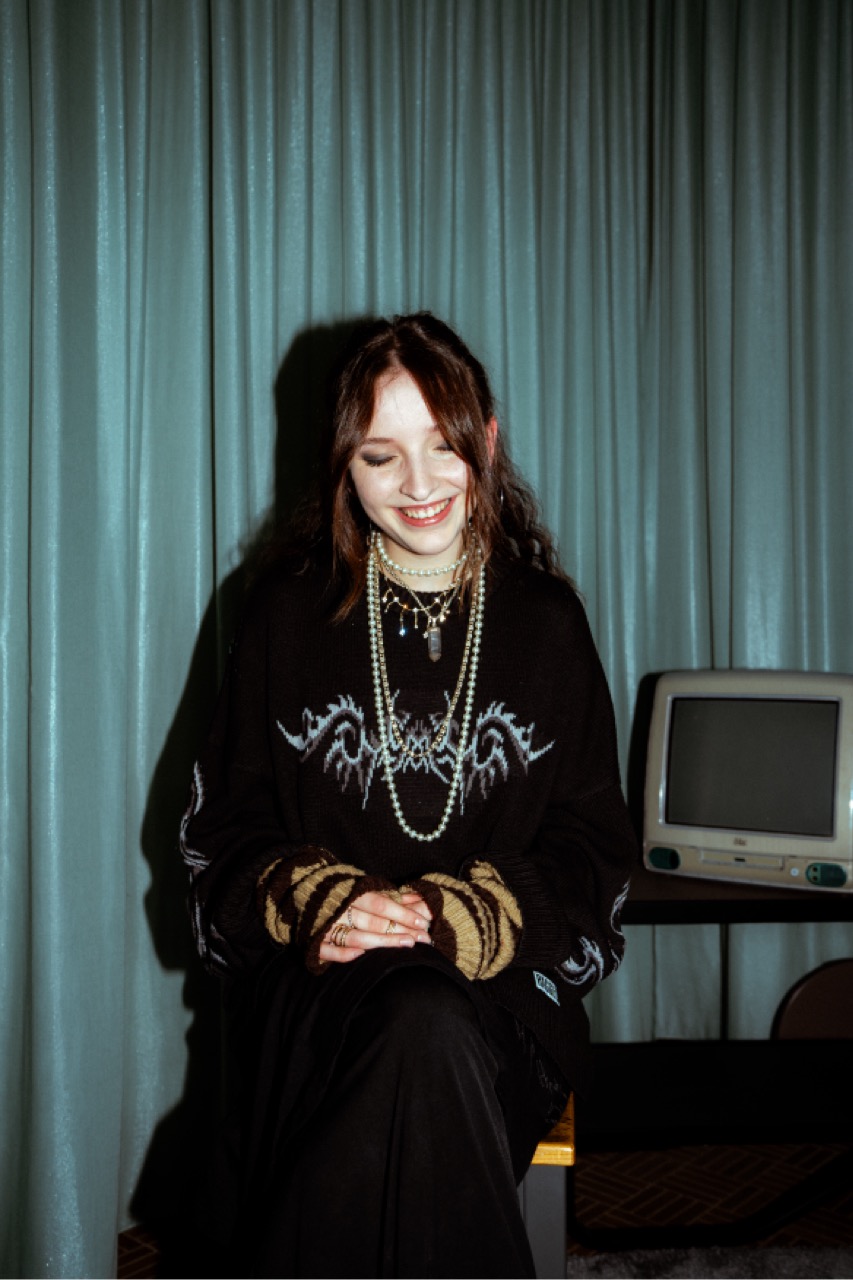 Erin lachend im dunklen Pullover mit Tribal-Print und mehreren Perlenketten.