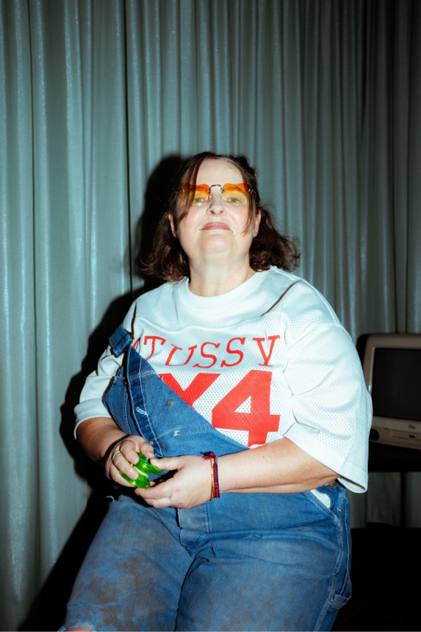 Katrin in Jeans-Latzhose und Trikot, mit getönter Brille und einem grünen Spielzeug in der Hand.