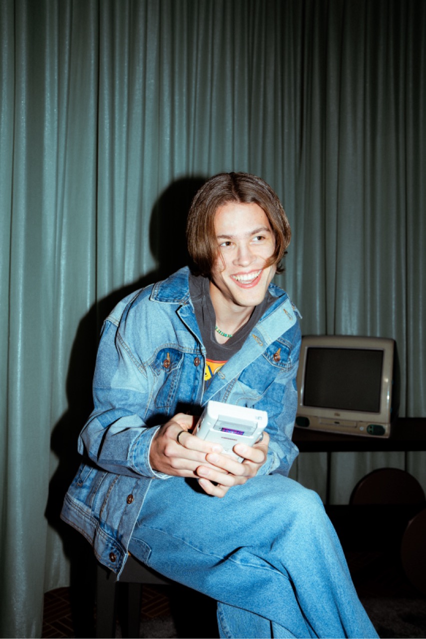 Joel im Jeans-Outfit, lachend mit einer alten Handheld-Spielkonsole in der Hand.