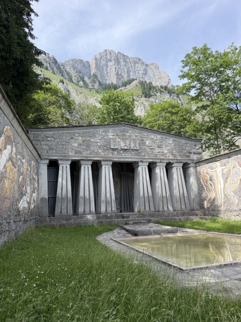 Steinerner Tempel mit Säulen und Wandmalereien vor einer Bergkulisse, davor ein Wasserbecken.