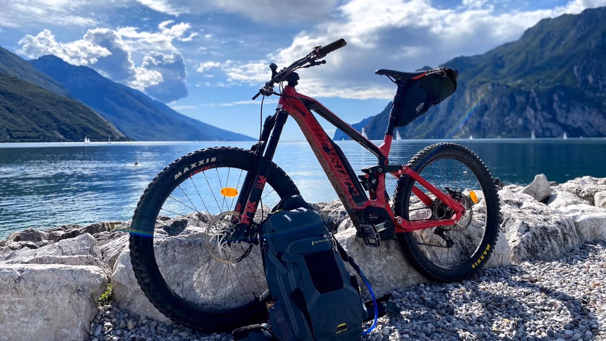 Mountainbike an einem Bergsee-Ufer, im Hintergrund Segelboote und Berge.