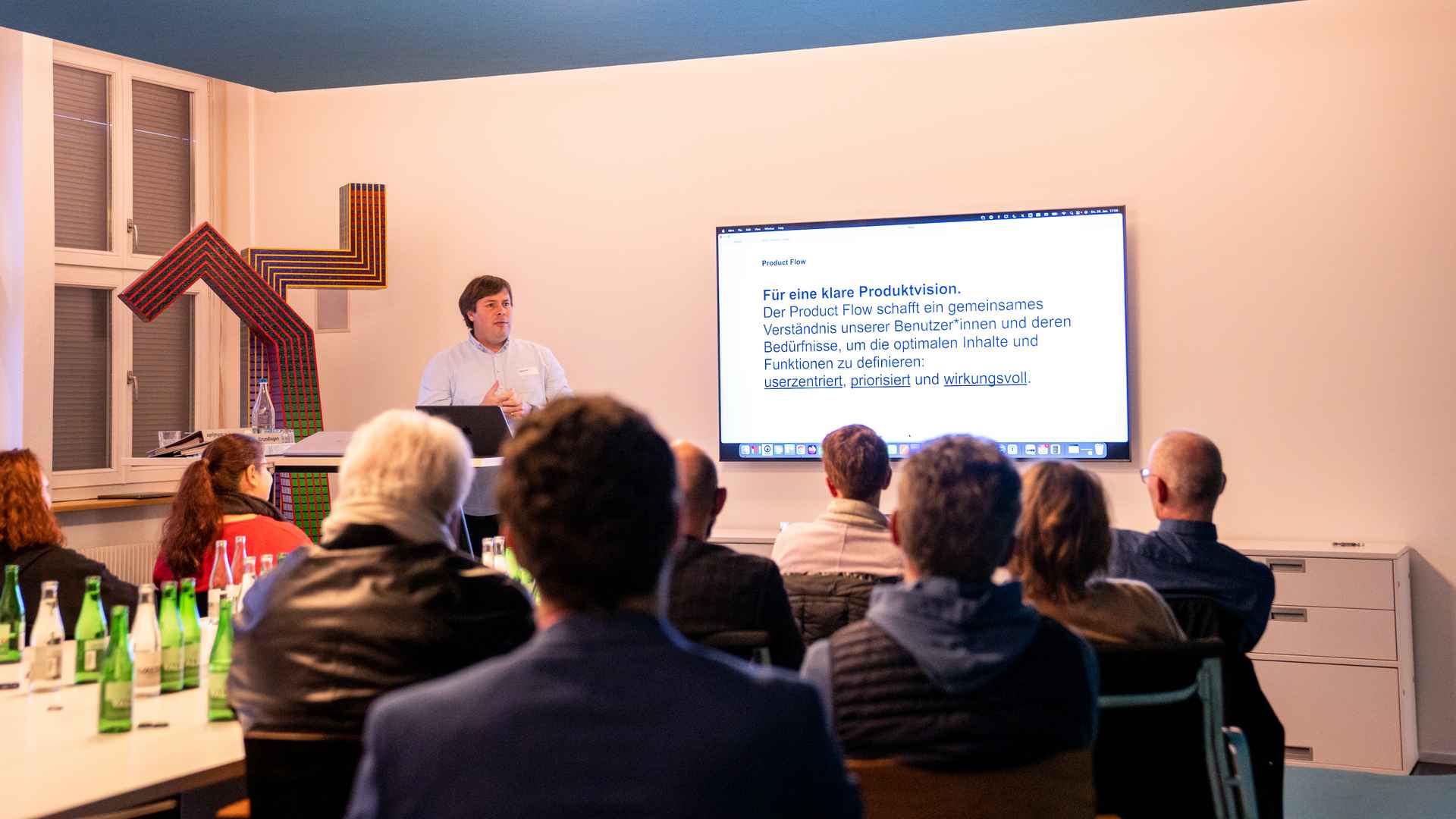 Referent am Laptop vor Publikum in einem Sitzungsraum, auf dem Bildschirm eine Folie mit dem Titel «Für eine klare Produktvision».