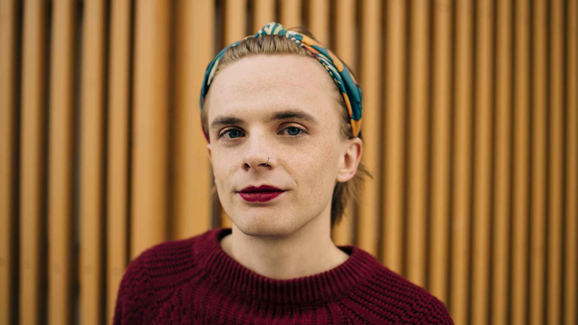 Porträt einer Person mit gemustertem Haarband, rotem Lippenstift und weinrotem Strickpullover vor einer Wand aus Holzlatten.