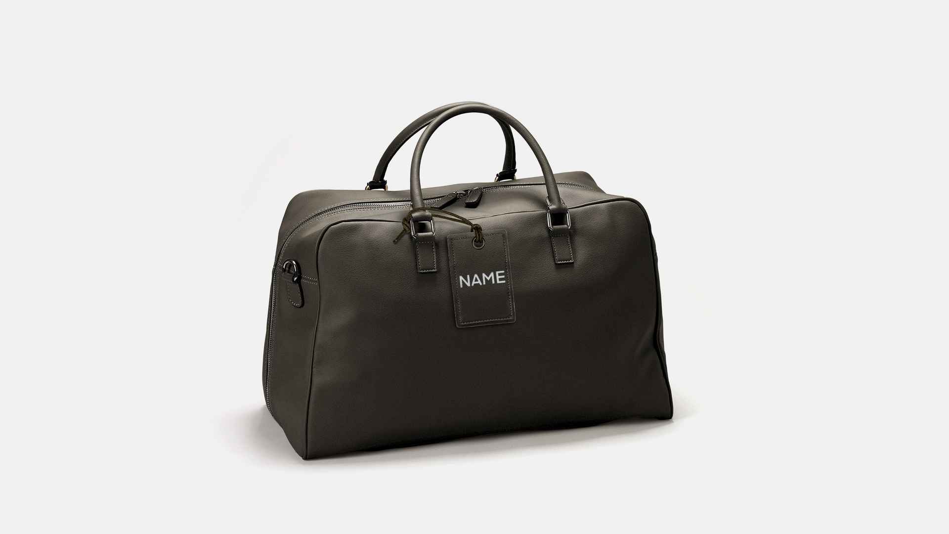 Dunkle Reisetasche aus Leder mit Gepäckanhänger, auf dem in weisser Schrift «NAME» steht, vor hellem Hintergrund.