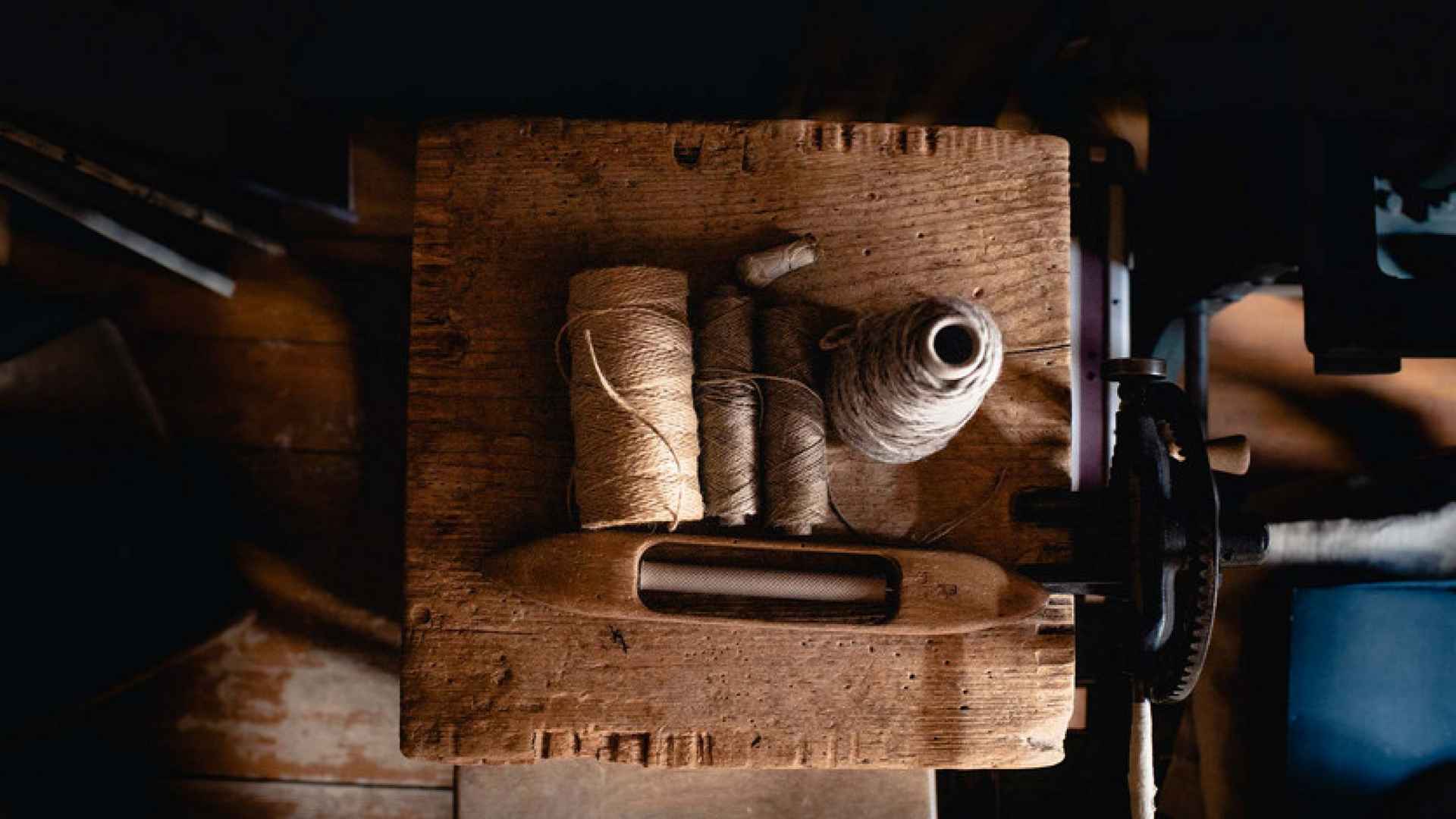 Aufsicht auf einen alten Holzwebstuhl mit mehreren Garnspulen aus Naturfaser und einem hölzernen Webschiffchen, dunkel und stimmungsvoll beleuchtet.