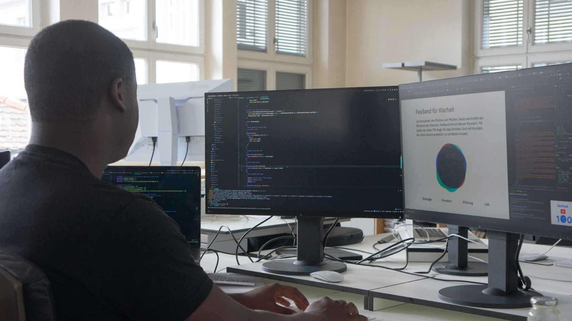 Mann sitzt von hinten gesehen an einem Arbeitsplatz mit mehreren Bildschirmen voller Programmcode und einer Festland-Webseite.