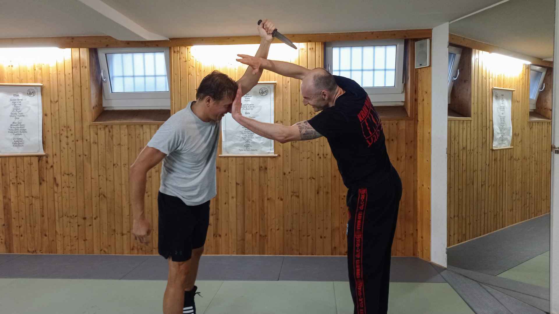 Zwei Männer demonstrieren in einem holzverkleideten Trainingsraum eine Krav-Maga-Abwehr gegen einen Messerangriff von oben.