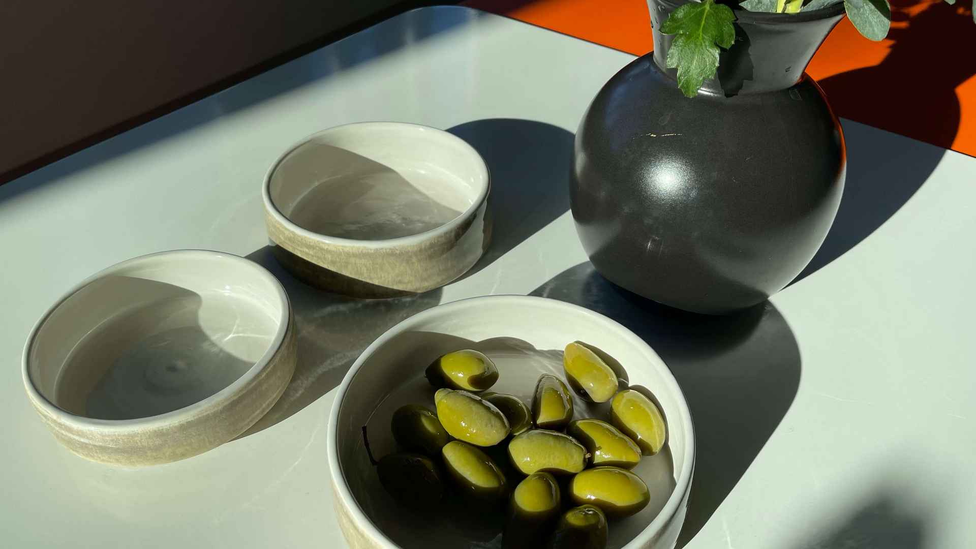 Handgemachte Keramikschalen und eine dunkle Vase mit Efeu auf einem sonnigen Tisch, in einer Schale grüne Oliven.