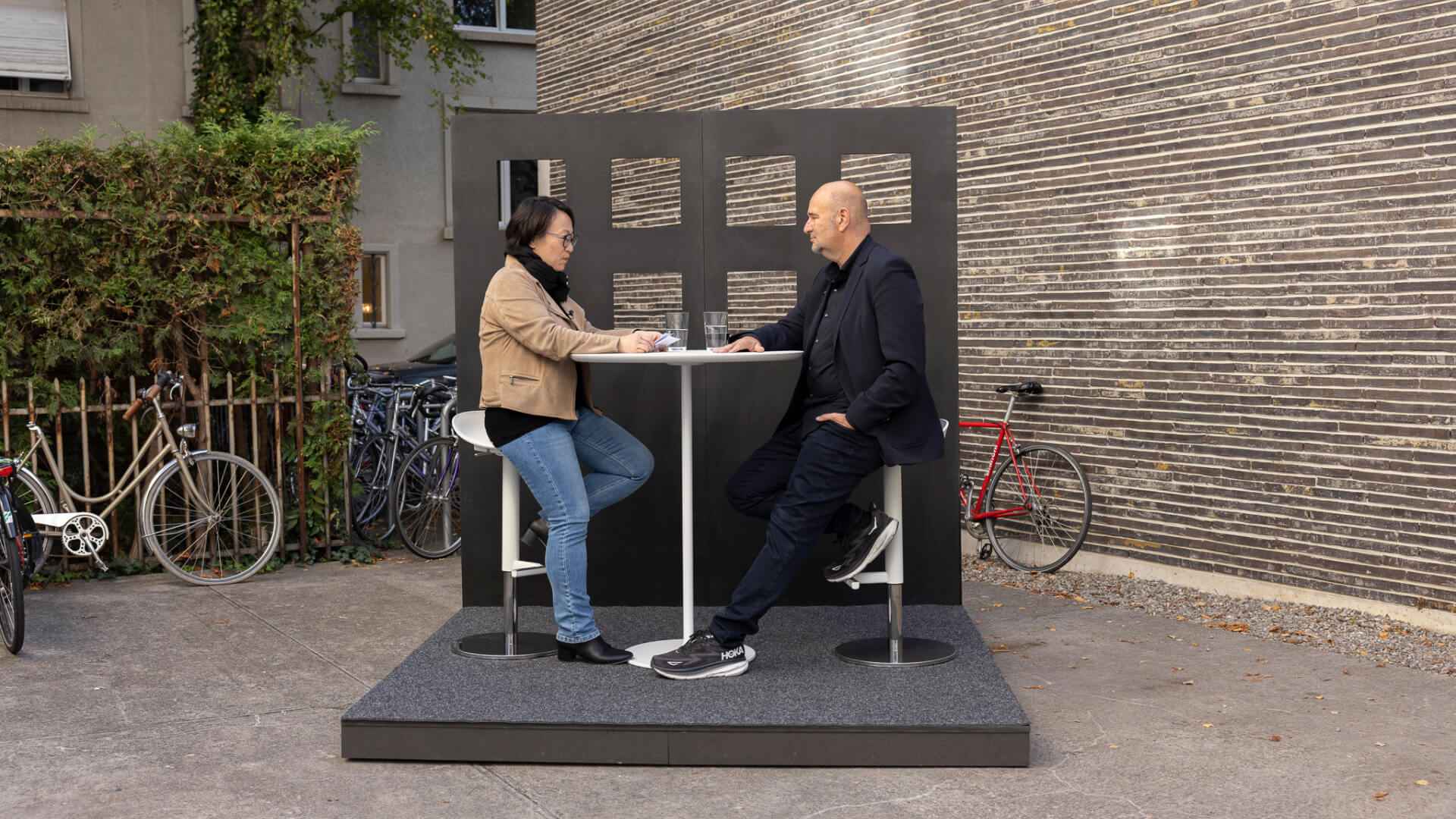 Frau und Mann sitzen sich an einem Stehtisch auf einem Podest im Freien gegenüber, vor einer Backsteinwand und abgestellten Velos.