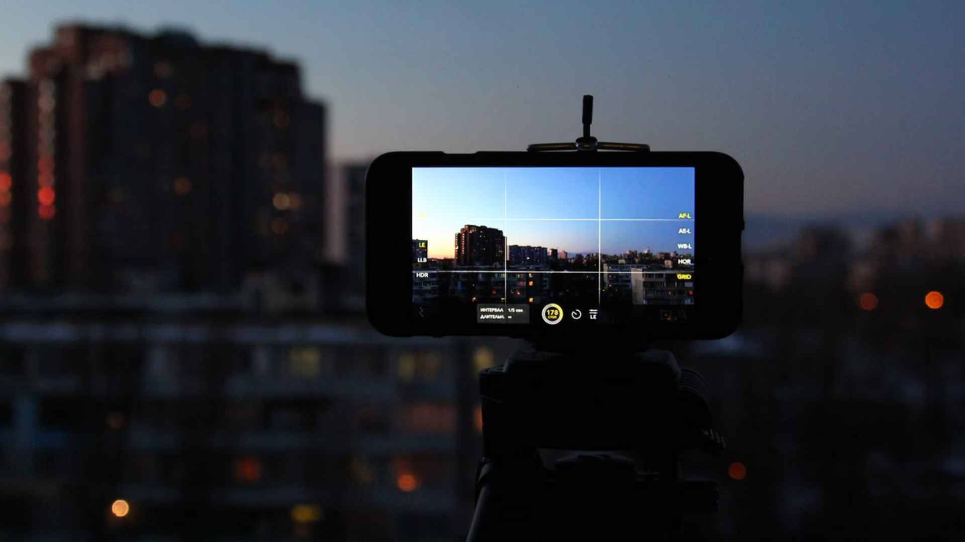 Smartphone auf einem Stativ filmt in der Abenddämmerung eine Stadtsilhouette mit beleuchteten Hochhäusern, das Display zeigt die Kamera-App.