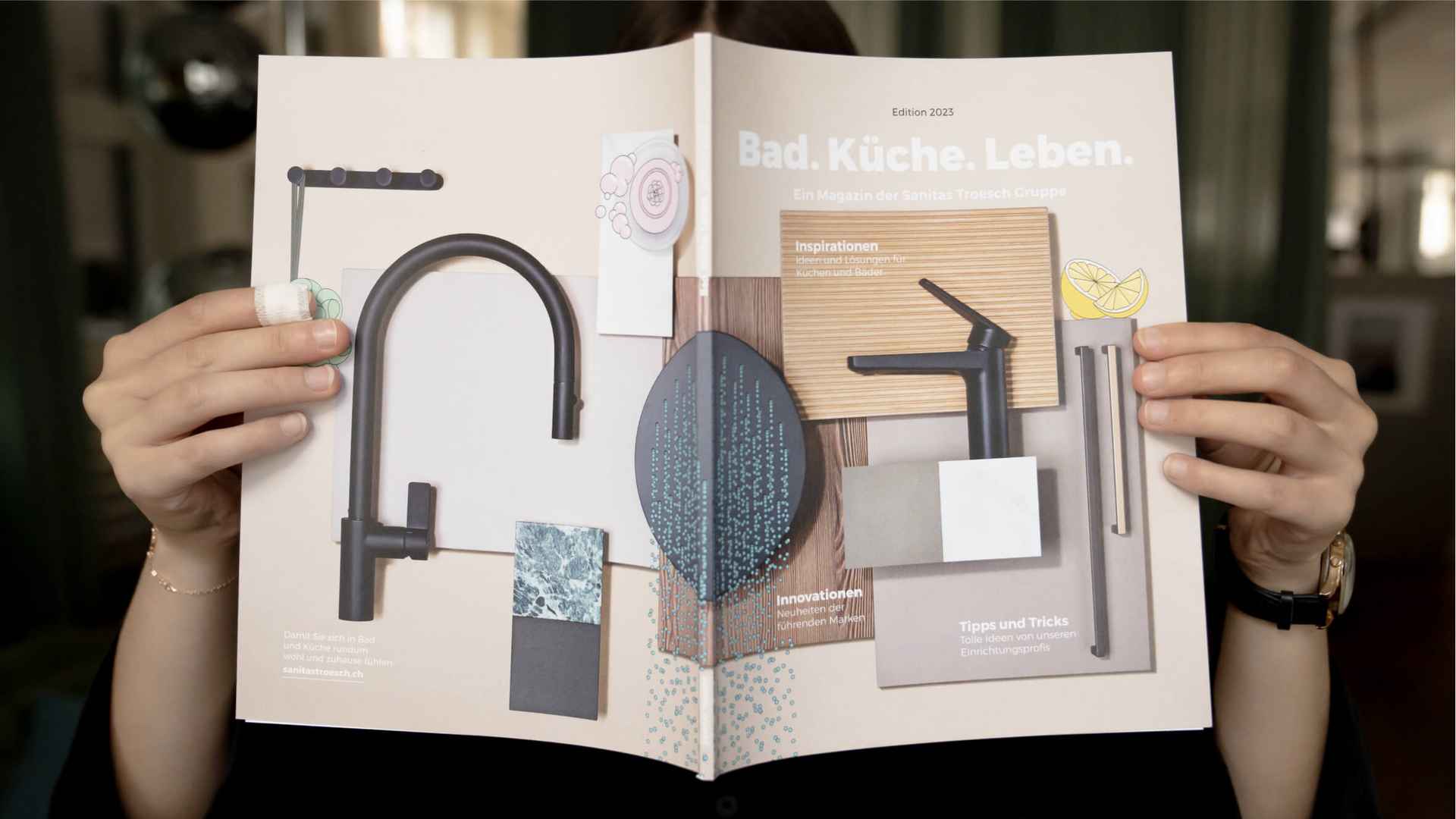 Person hält das aufgeschlagene Magazin «Bad. Küche. Leben.» vors Gesicht, das Cover zeigt Armaturen und Materialmuster in Beige und Anthrazit.