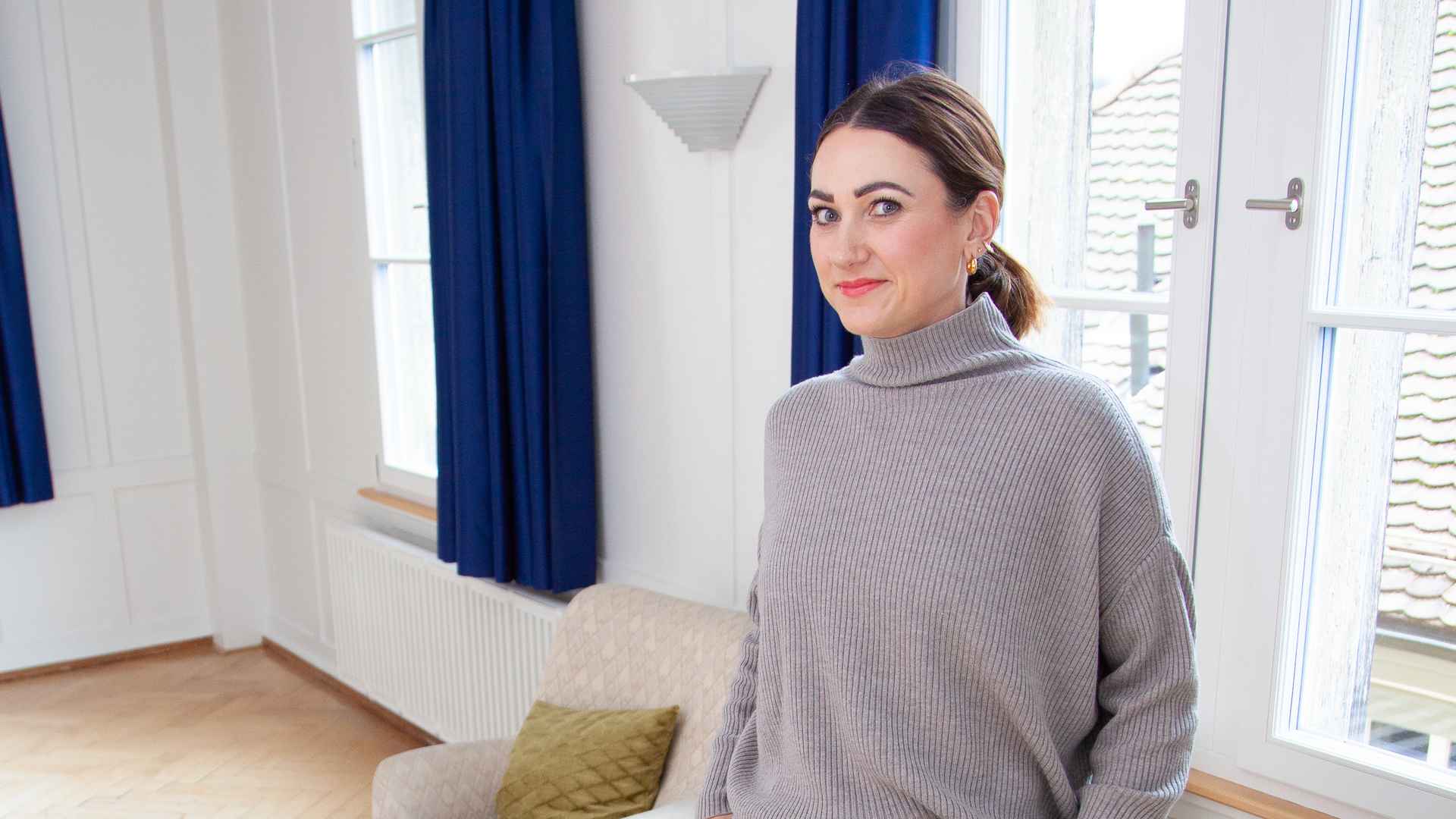 Lucia Burtscher im grauen Rollkragenpullover in einem hellen Raum mit dunkelblauen Vorhängen, Sofa und Blick über die Dächer.