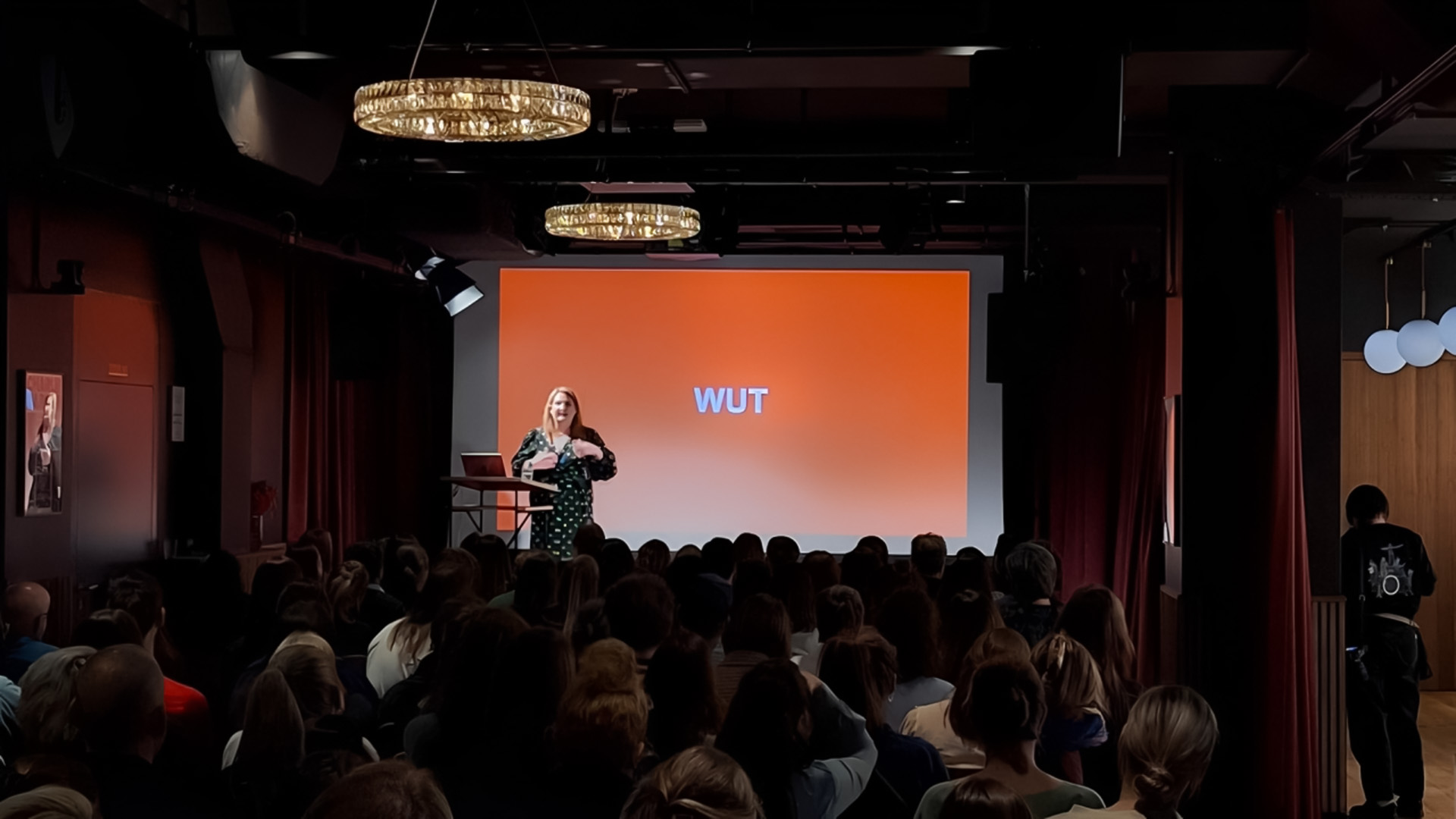 Eine Rednerin steht auf einer Bühne vor einer orangen Leinwand mit dem Wort «WUT», davor ein voll besetzter, dunkler Saal.