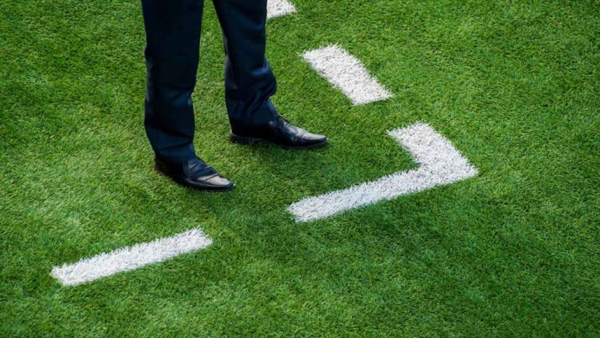 Beine einer Person in Anzughose und schwarzen Lederschuhen auf Kunstrasen mit weissen Spielfeldmarkierungen.