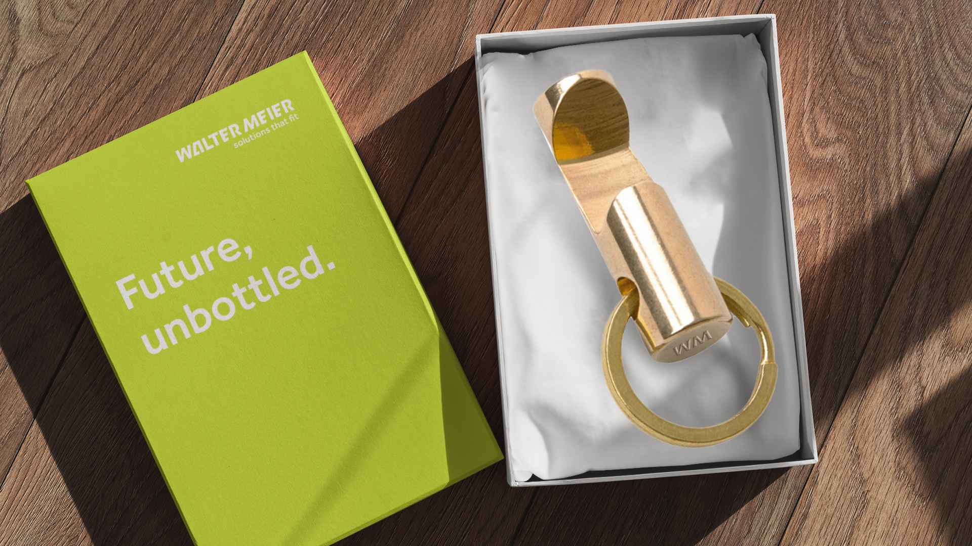 Goldener Flaschenöffner mit Schlüsselring in weisser Geschenkbox neben grüner Walter-Meier-Karte mit «Future, unbottled.» auf Holzboden.