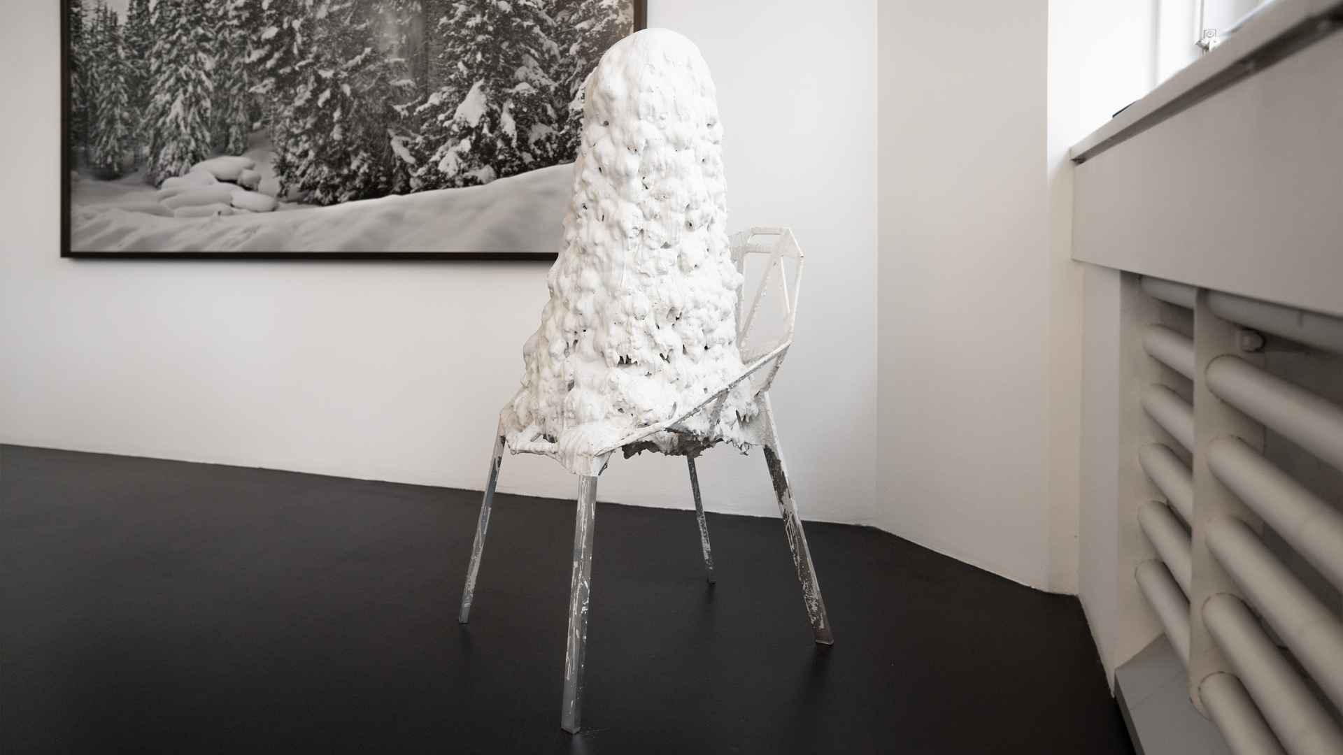 Skulptur von Beni Bischof: Metallstuhl mit hoher, weisser, wulstiger Masse vor gerahmtem Schwarzweissfoto eines verschneiten Waldes.