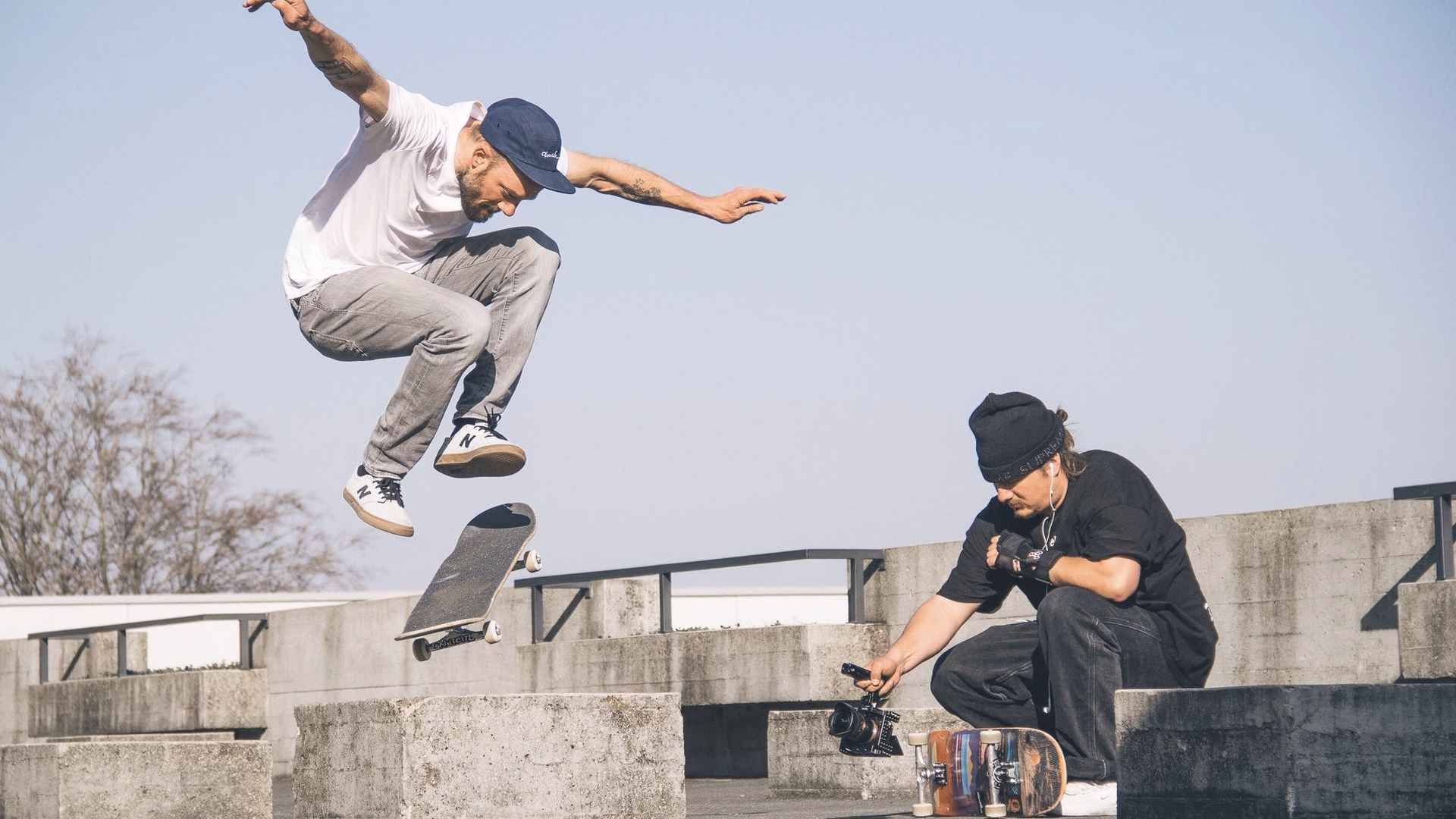 Skater mit Mütze springt einen Ollie über Betonblöcke, daneben kauert ein Mann in Schwarz und filmt mit einer Kamera.