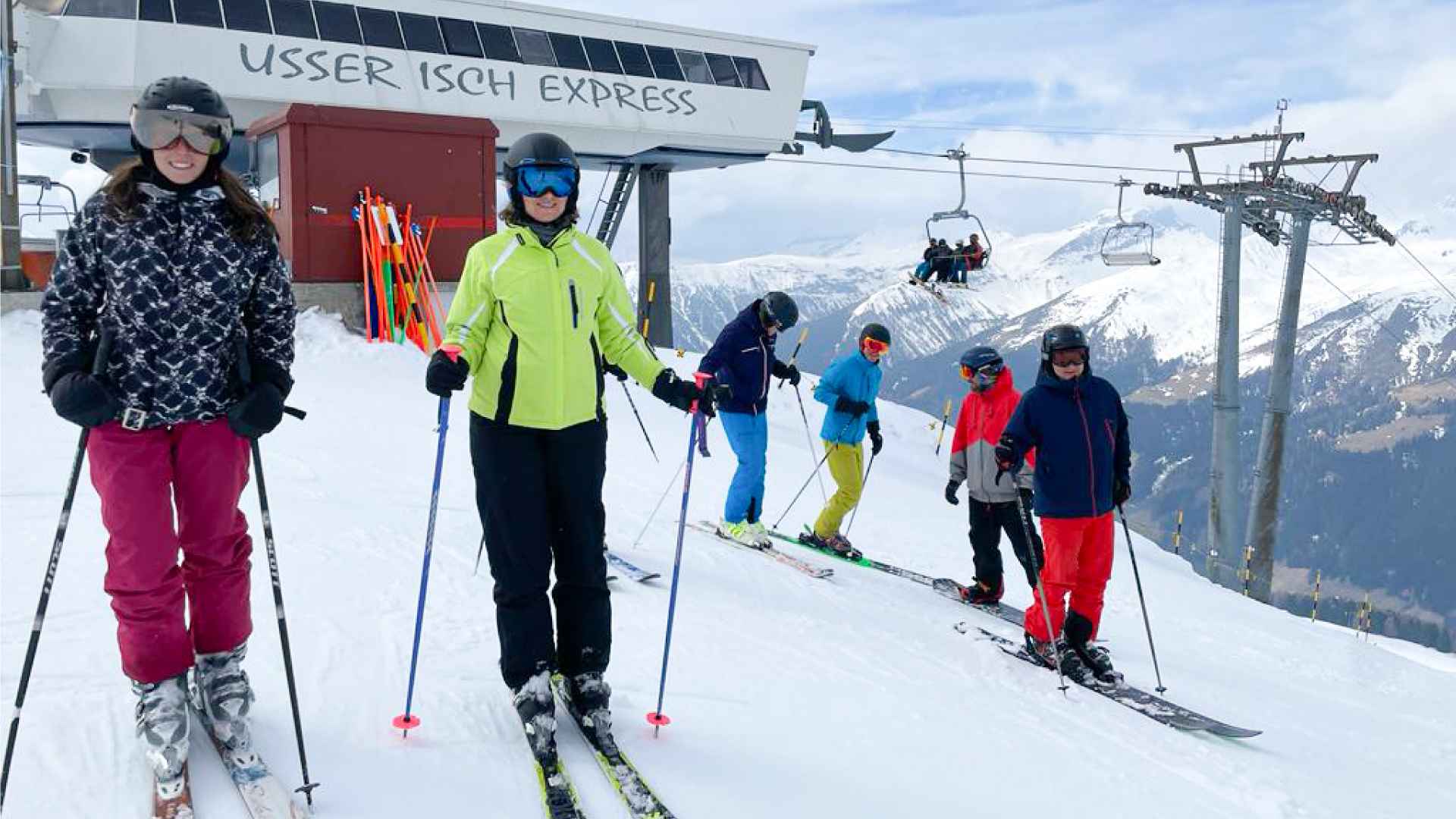 Skifahrende in bunter Ausrüstung bei der Bergstation «Usser Isch Express», dahinter Sessellift und verschneite Berge.