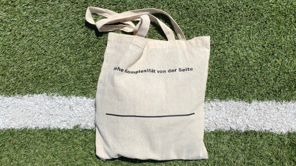 Stofftasche mit dem Aufdruck «hohe Komplexität von der Seite» liegt auf Kunstrasen neben einer weissen Linie.