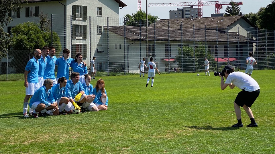 Fotografin in Weiss und Schwarz fotografiert das Festland-Team in Hellblau am Spielfeldrand, im Hintergrund spielen Teams in Rot und Weiss