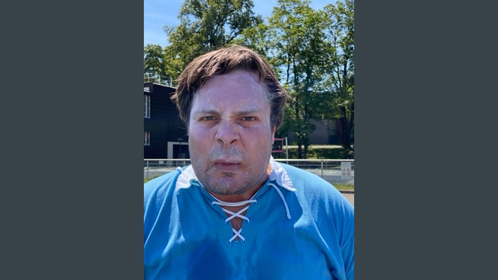 Spieler im hellblauen Retro-Trikot mit Schnürkragen blickt ernst in die Kamera, im Hintergrund Bäume und der Sportplatz bei Sonnenschein