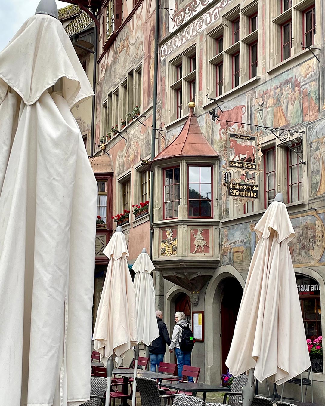 Bemalte Altstadtfassaden mit Erker und Wirtshausschild mit rotem Ochsen, davor geschlossene beige Sonnenschirme