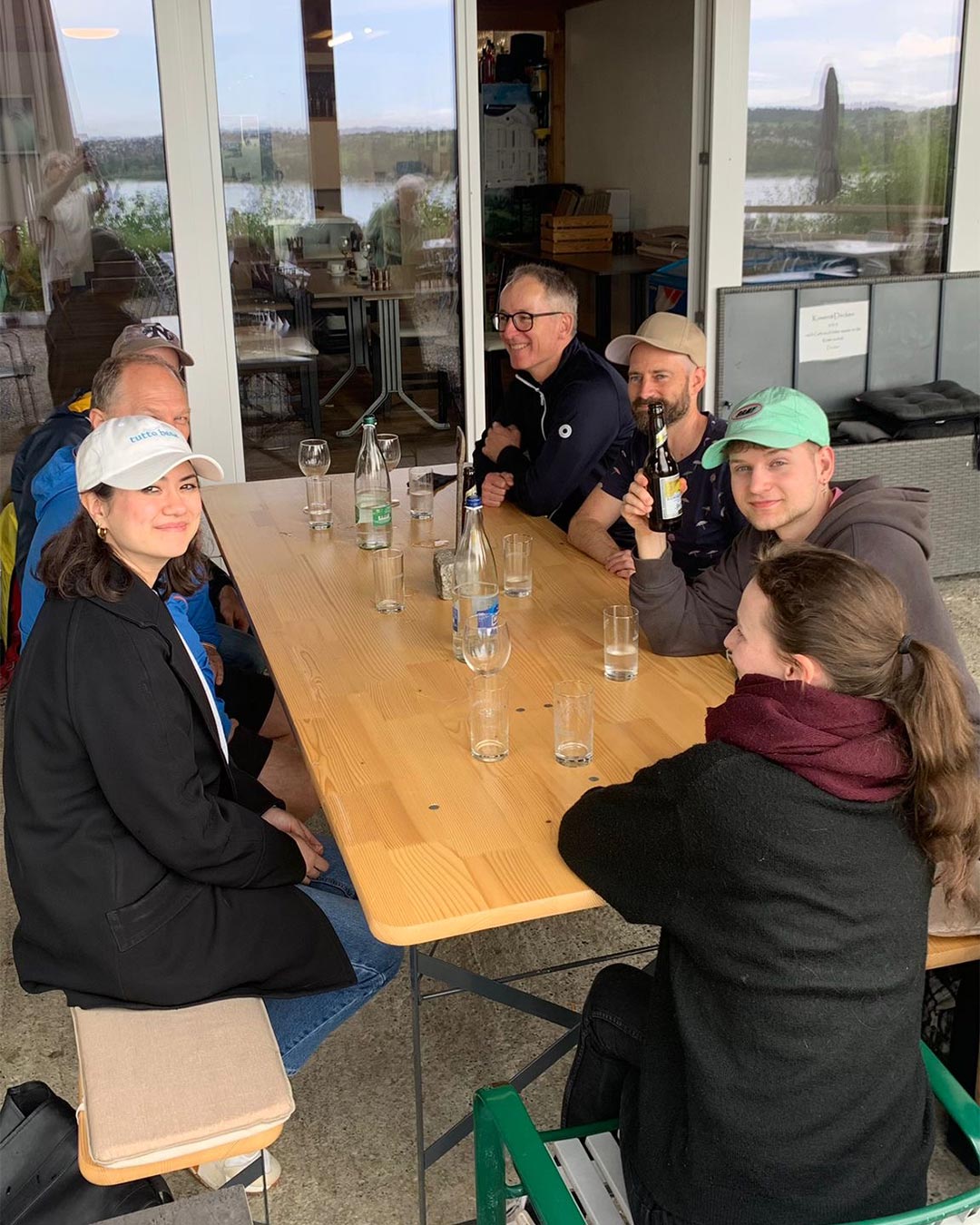 Team in Mützen und Jacken an einem langen Holztisch auf einer Terrasse mit Getränken, im Hintergrund ein See