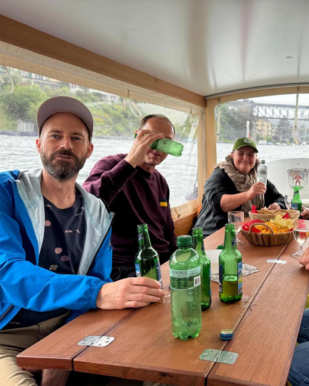 Drei Personen an einem Holztisch in einem überdachten Boot auf dem Rhein, mit Mineralwasser und Chips, einer trinkt aus der Flasche