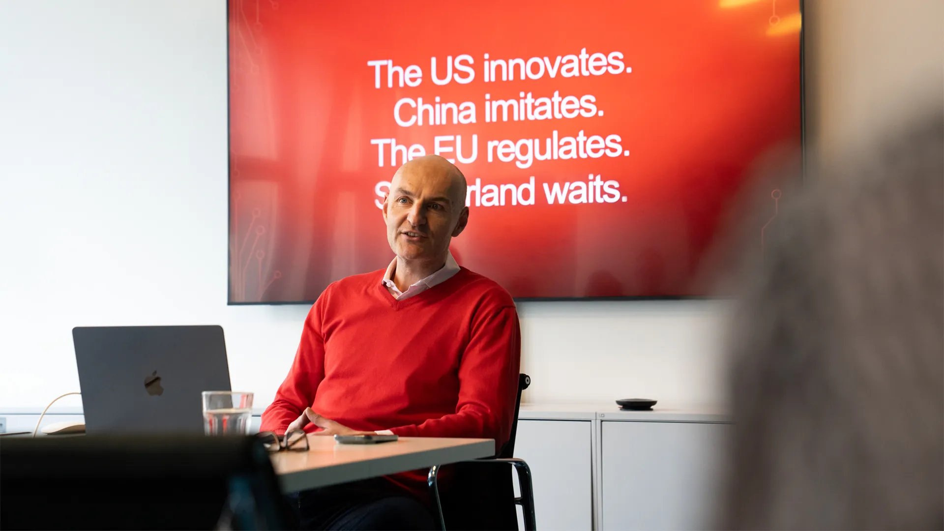Christian Sidow im roten Pullover referiert vor einem Bildschirm mit dem Text «The US innovates. China imitates. The EU regulates.»