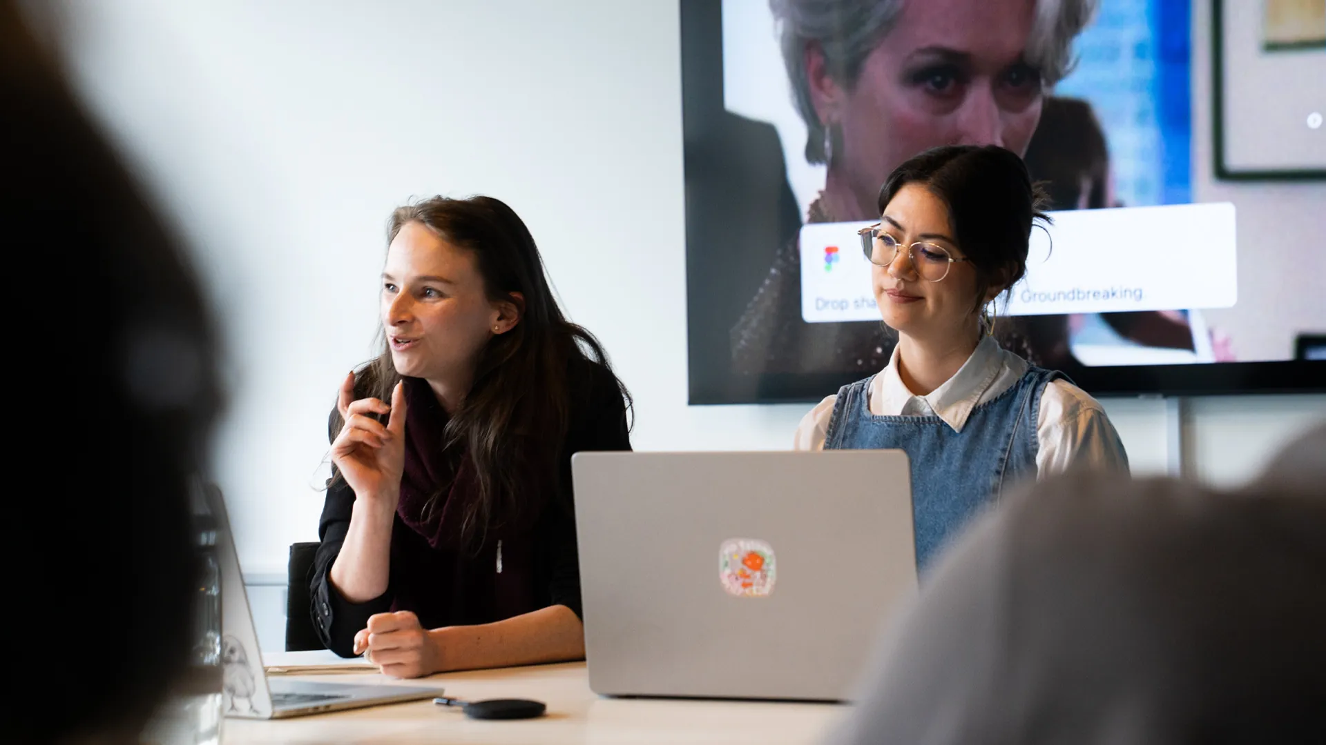 Zwei Frauen sitzen mit Laptop an einem Tisch und sprechen zum Publikum, hinter ihnen zeigt ein grosser Bildschirm eine Filmszene.