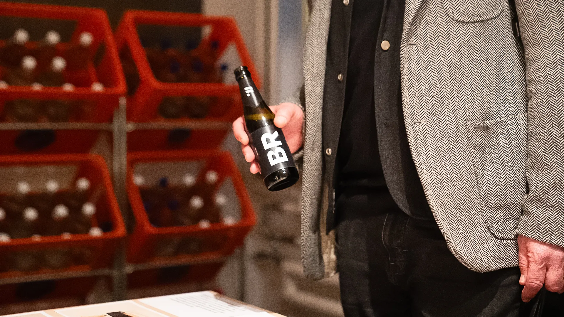 Mann im grauen Fischgrat-Sakko hält eine dunkle Bierflasche mit grosser «BR»-Etikette, im Hintergrund gestapelte rote Getränkeharasse.