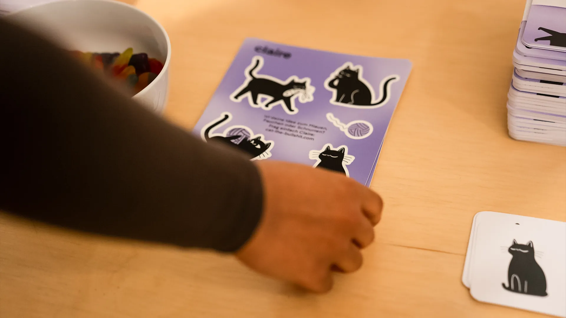 Hand greift nach einem violetten Stickerbogen «claire» mit schwarzen Katzen-Illustrationen, daneben Kartenstapel und eine Schale Fruchtgummis.