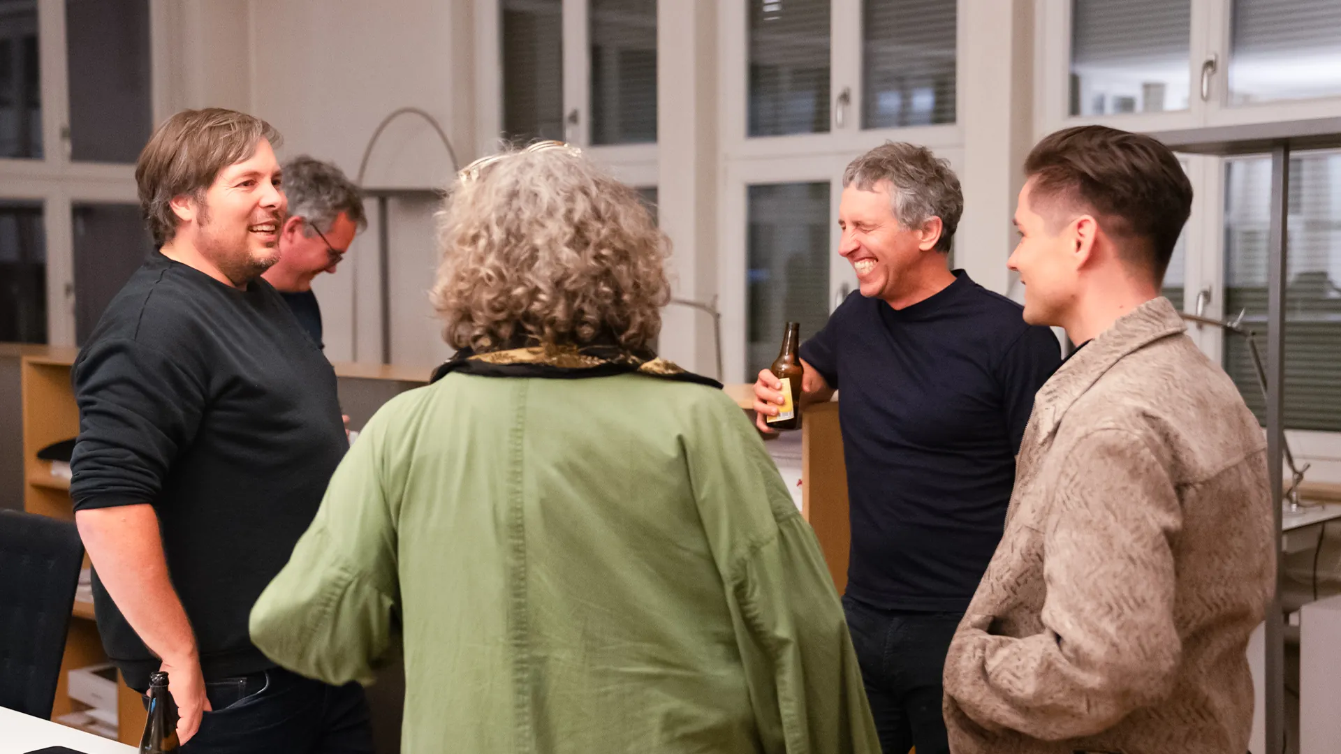 Gäste der Design Night stehen in einem hellen Büroraum in lockerer Runde zusammen und lachen, einer hält eine Bierflasche in der Hand.