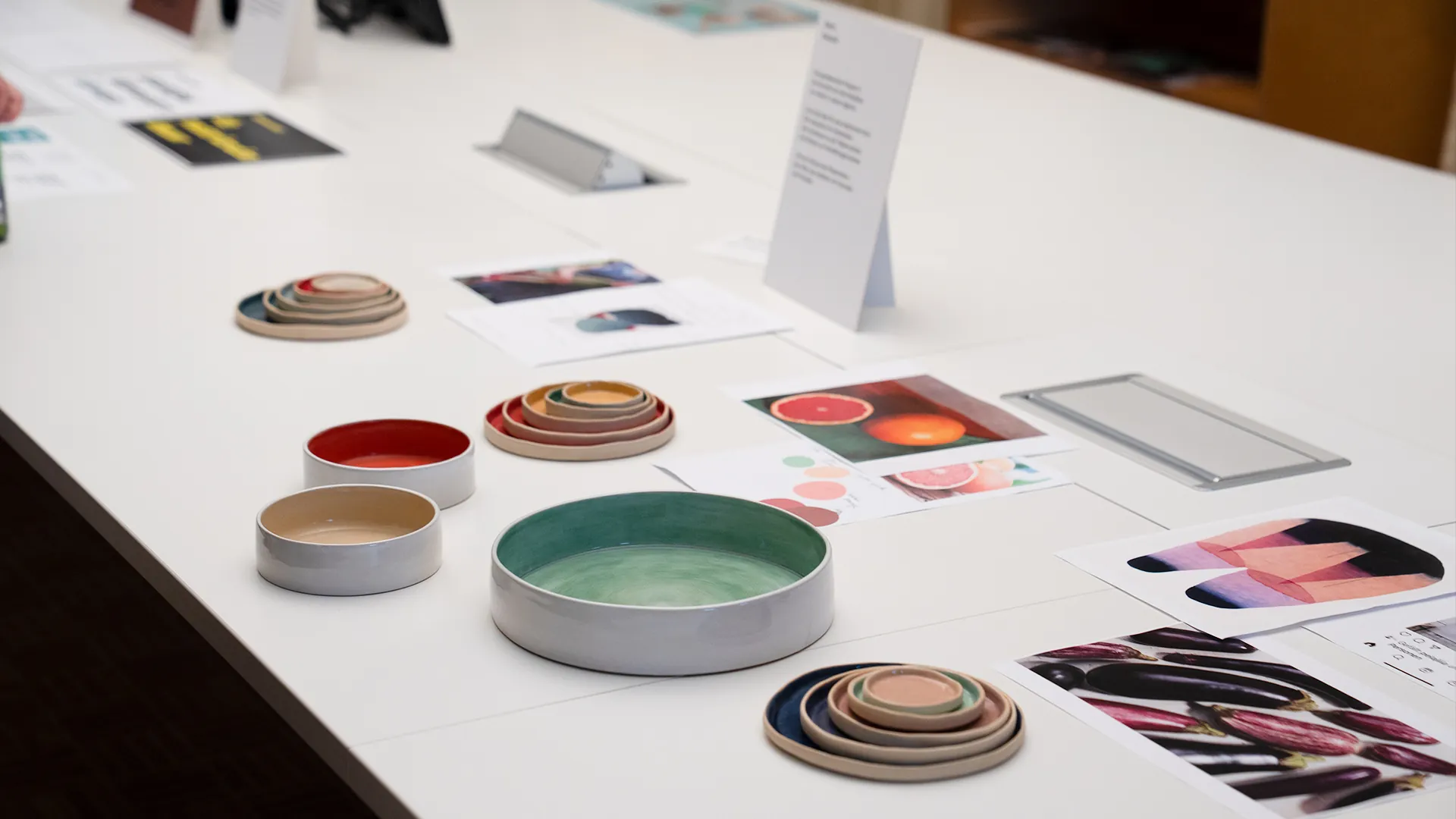 Ausstellungstisch mit gestapelten Keramikschalen in Rot, Grün und Pastelltönen, dazwischen farbige Fotografien von Früchten und Gemüse.