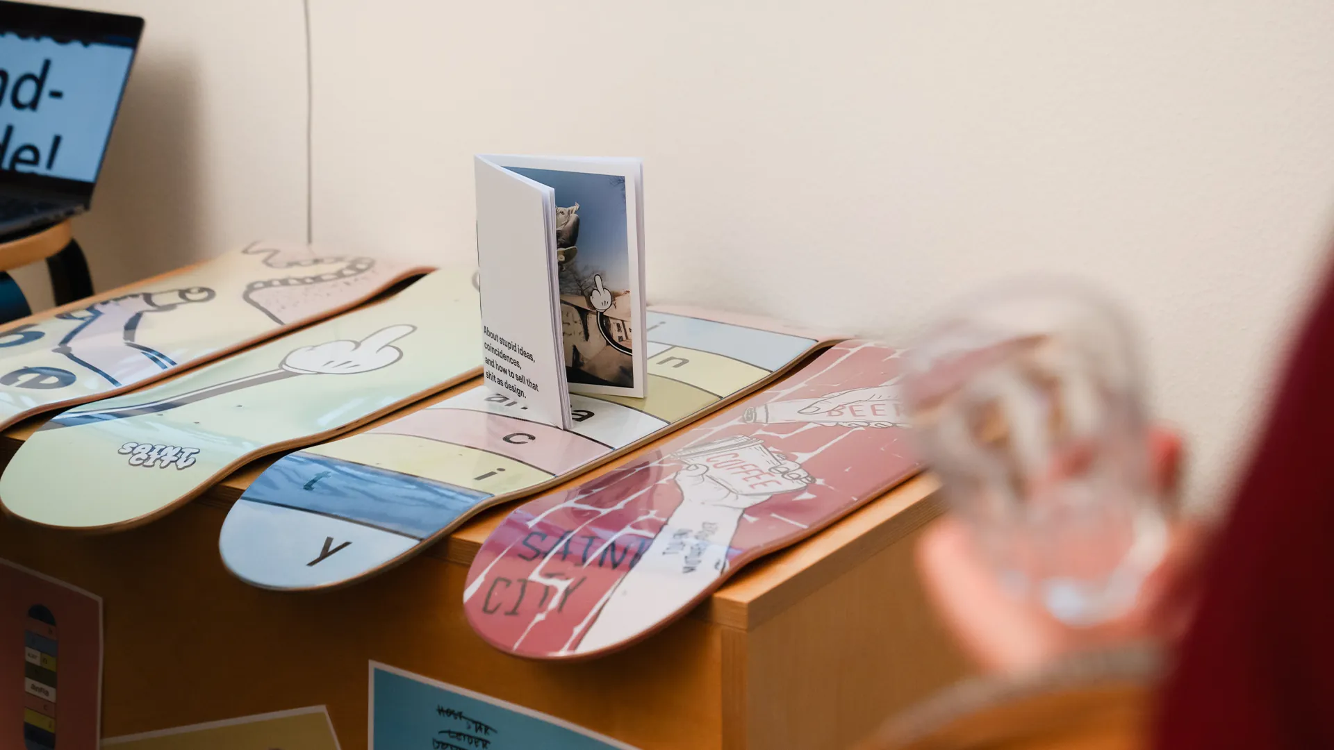 Mehrere bemalte Skateboard-Decks in Pastellfarben mit Comic-Illustrationen und einzelnen Buchstaben, dazu eine aufgestellte Broschüre.