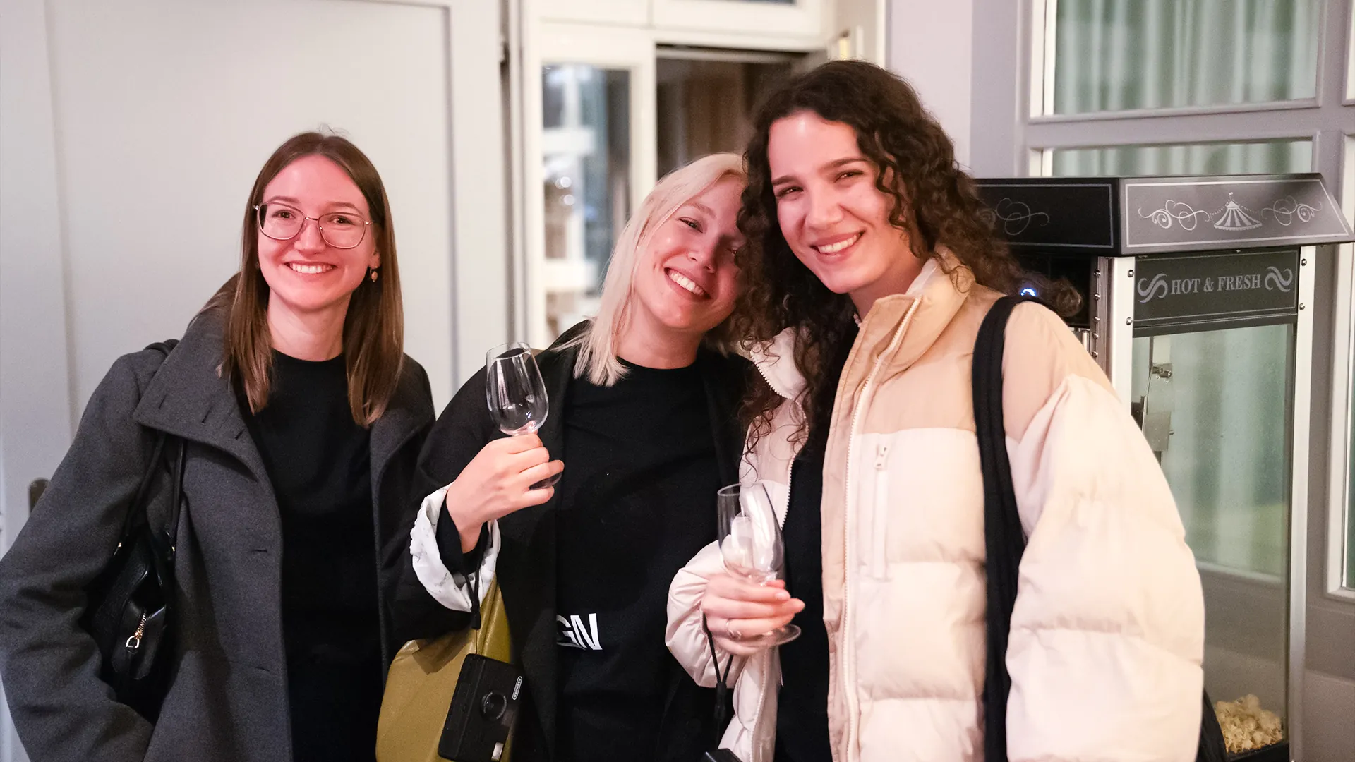 Drei Besucherinnen lachen mit Weingläsern in die Kamera, neben ihnen eine Popcornmaschine mit der Aufschrift «Hot & Fresh».
