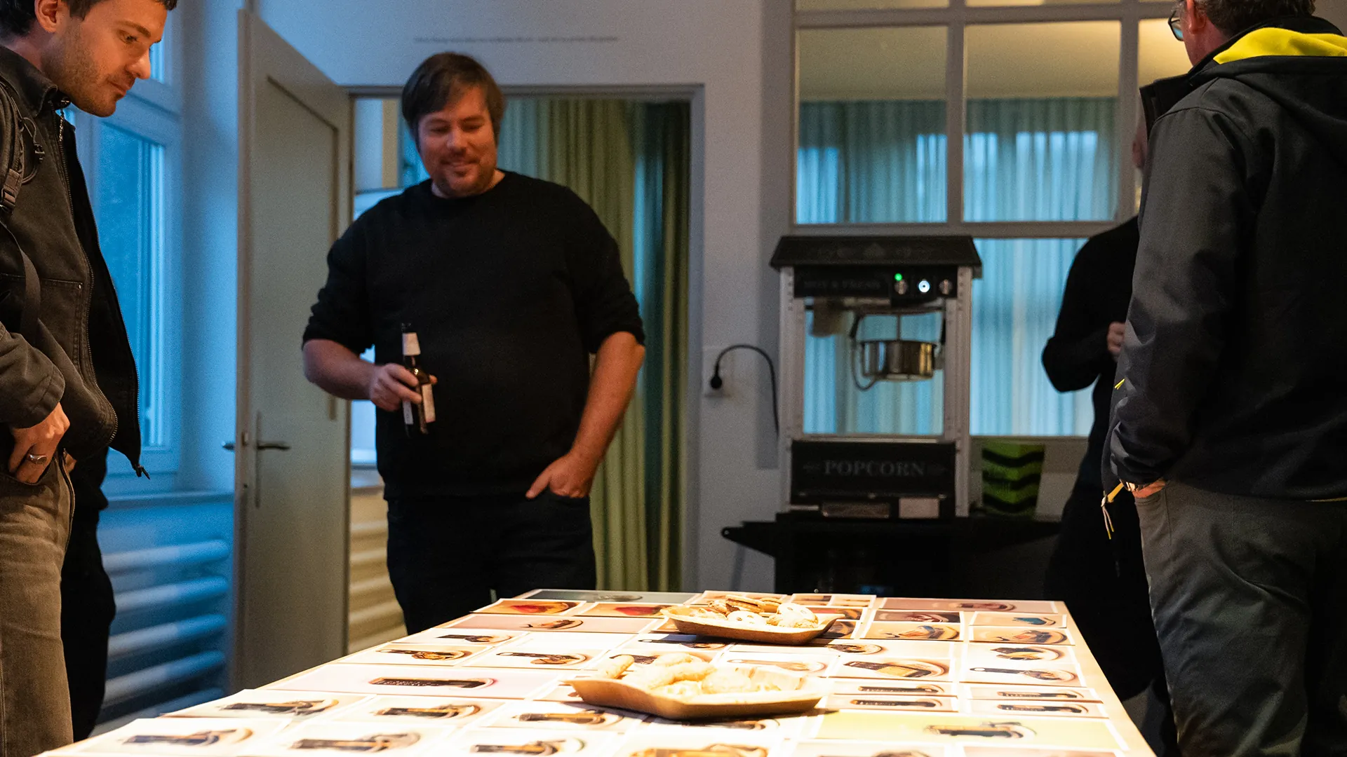 Gäste stehen um einen langen Tisch mit ausgelegten Karten und Snackschalen, im Hintergrund eine Popcornmaschine vor hohen Fenstern.