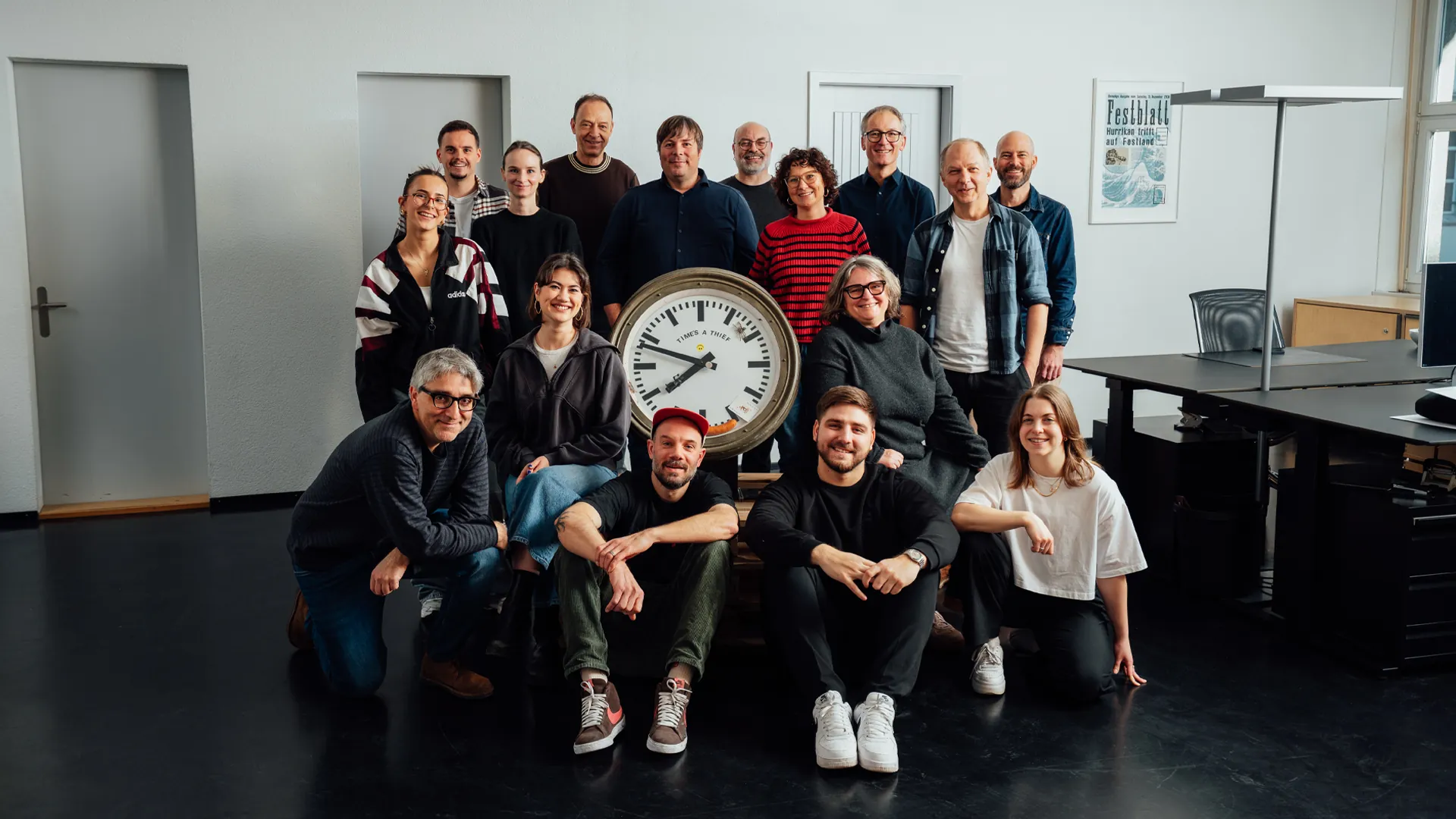 Das Festland-Team posiert in einem hellen Raum wie für ein Klassenfoto, in der Mitte hält eine Person eine grosse Bahnhofsuhr.