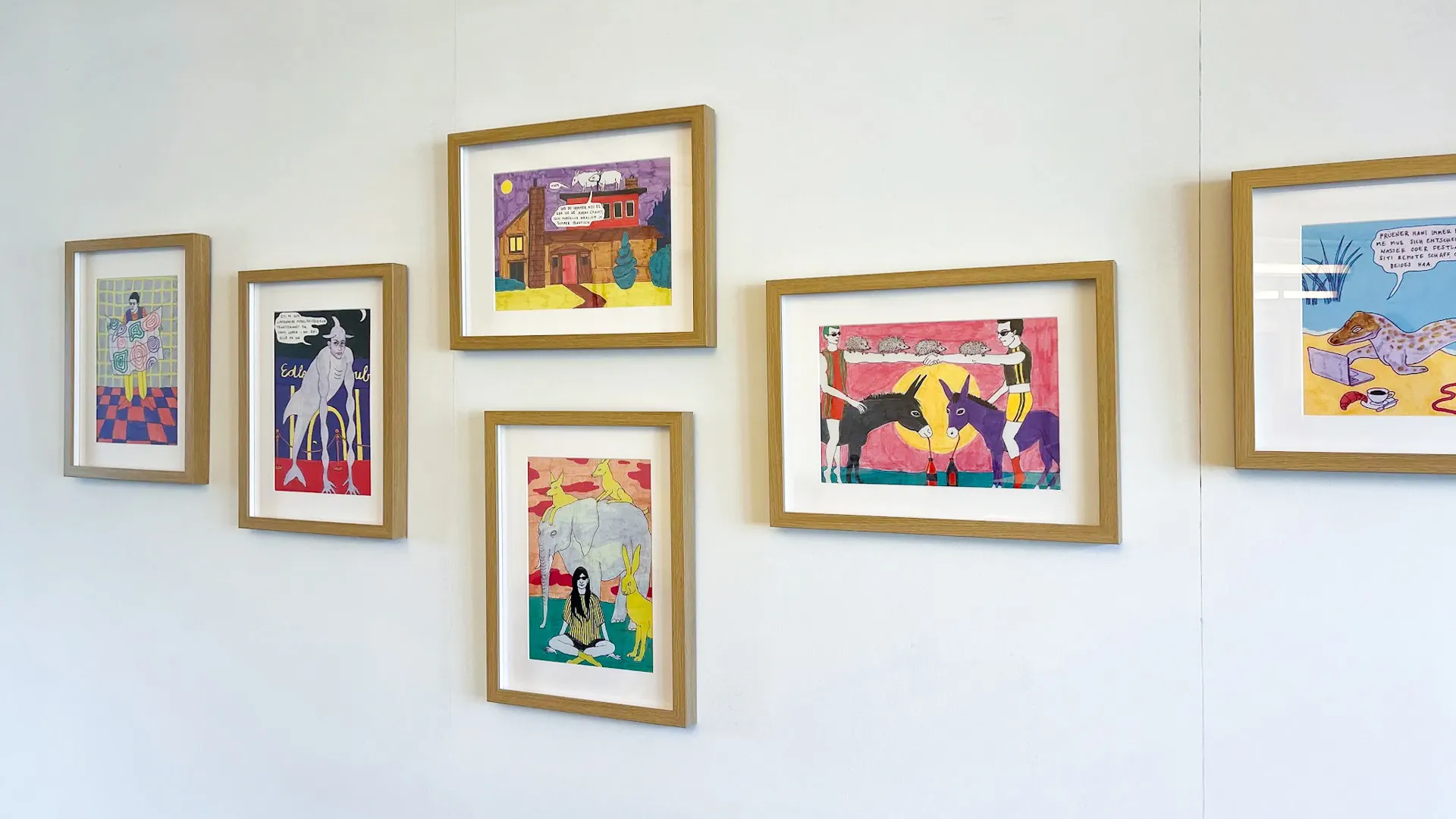 Gerahmte, farbenfrohe Comic-Illustrationen mit Figuren, Tieren und Sprechblasen hängen in lockerer Anordnung an einer weissen Wand.