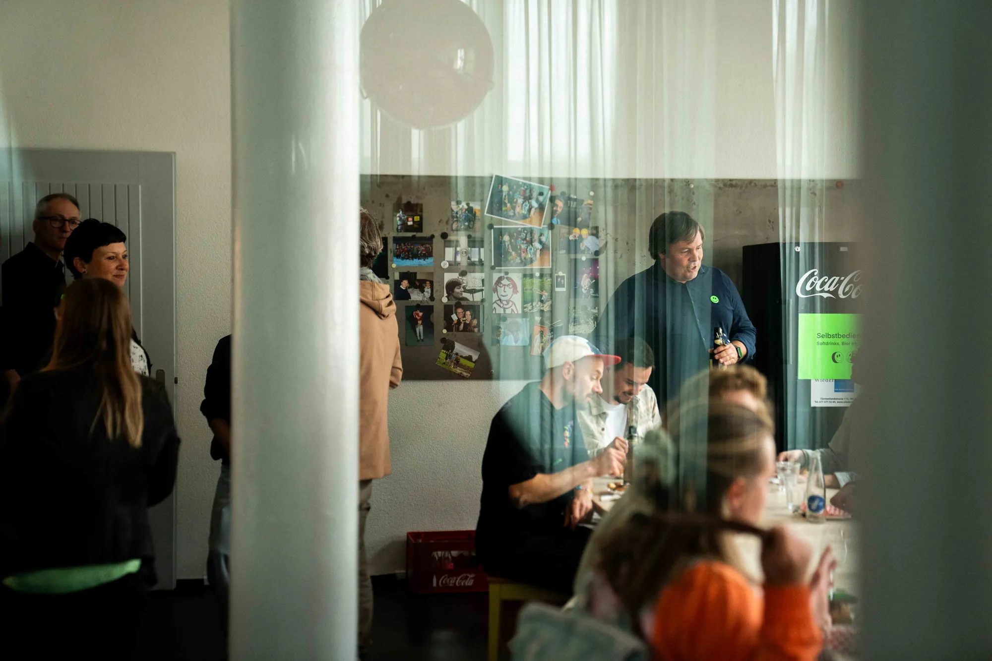 Blick durch einen transparenten Vorhang auf Gäste an einem Tisch, dahinter eine Wand voller Fotos und ein Coca-Cola-Kühlschrank.
