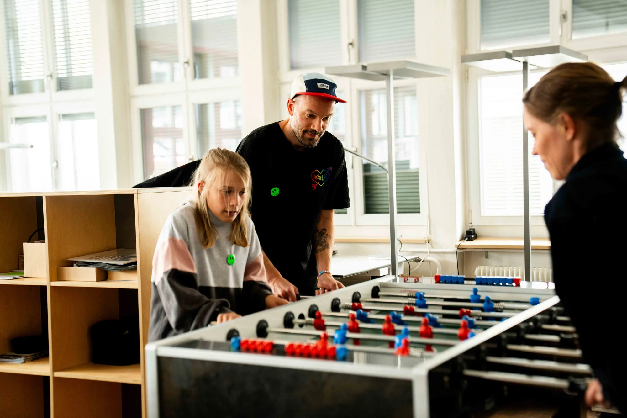 Ein Mädchen und ein Mann mit Schirmmütze spielen im hellen Agenturraum Tischfussball gegen eine Frau, alle mit grünen Smiley-Buttons.