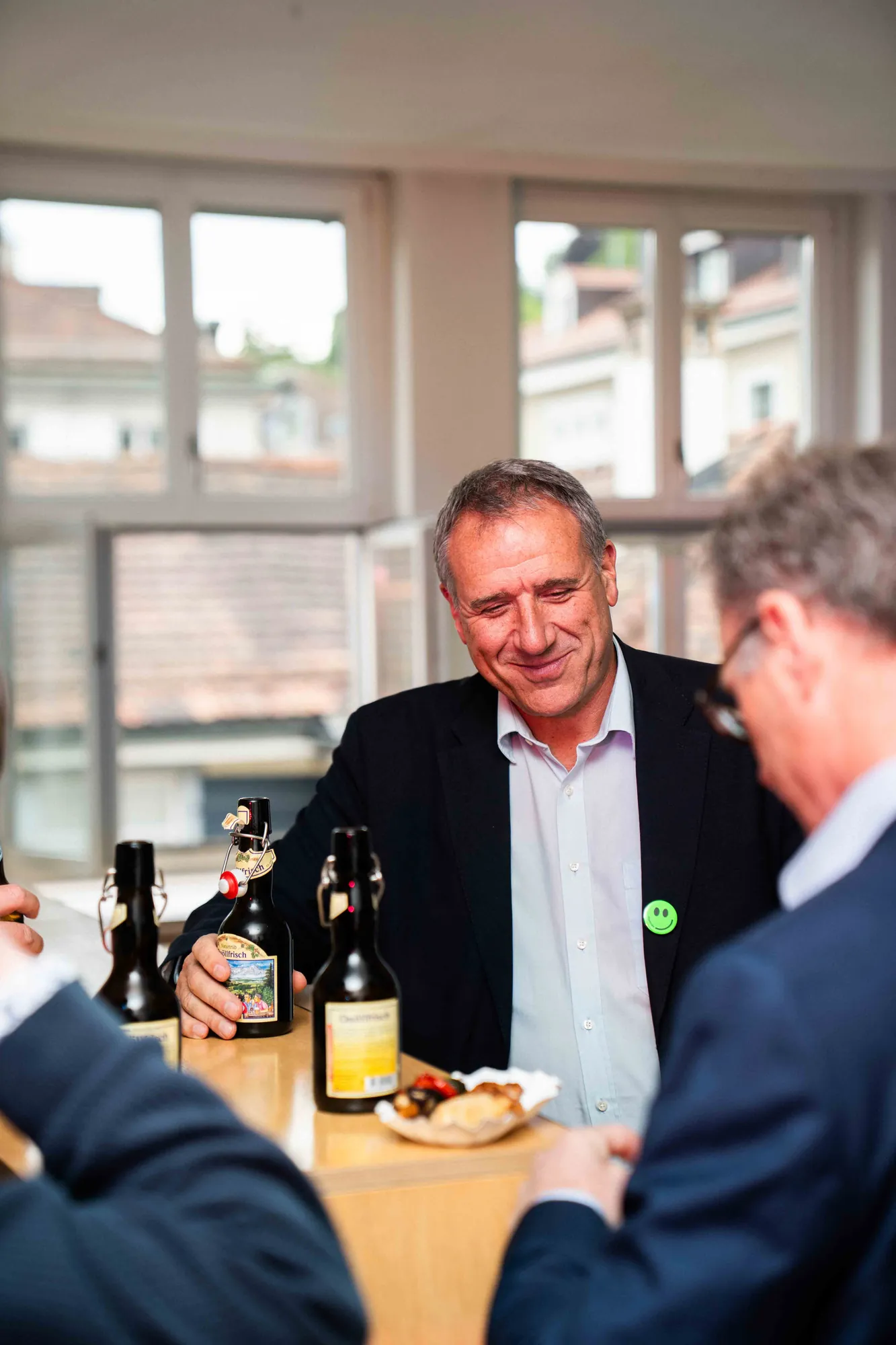 Lächelnder Mann im dunklen Sakko sitzt mit Bügelflaschen-Bier und einem Snack am Tisch, hinter ihm Fenster mit Blick über die Dächer.