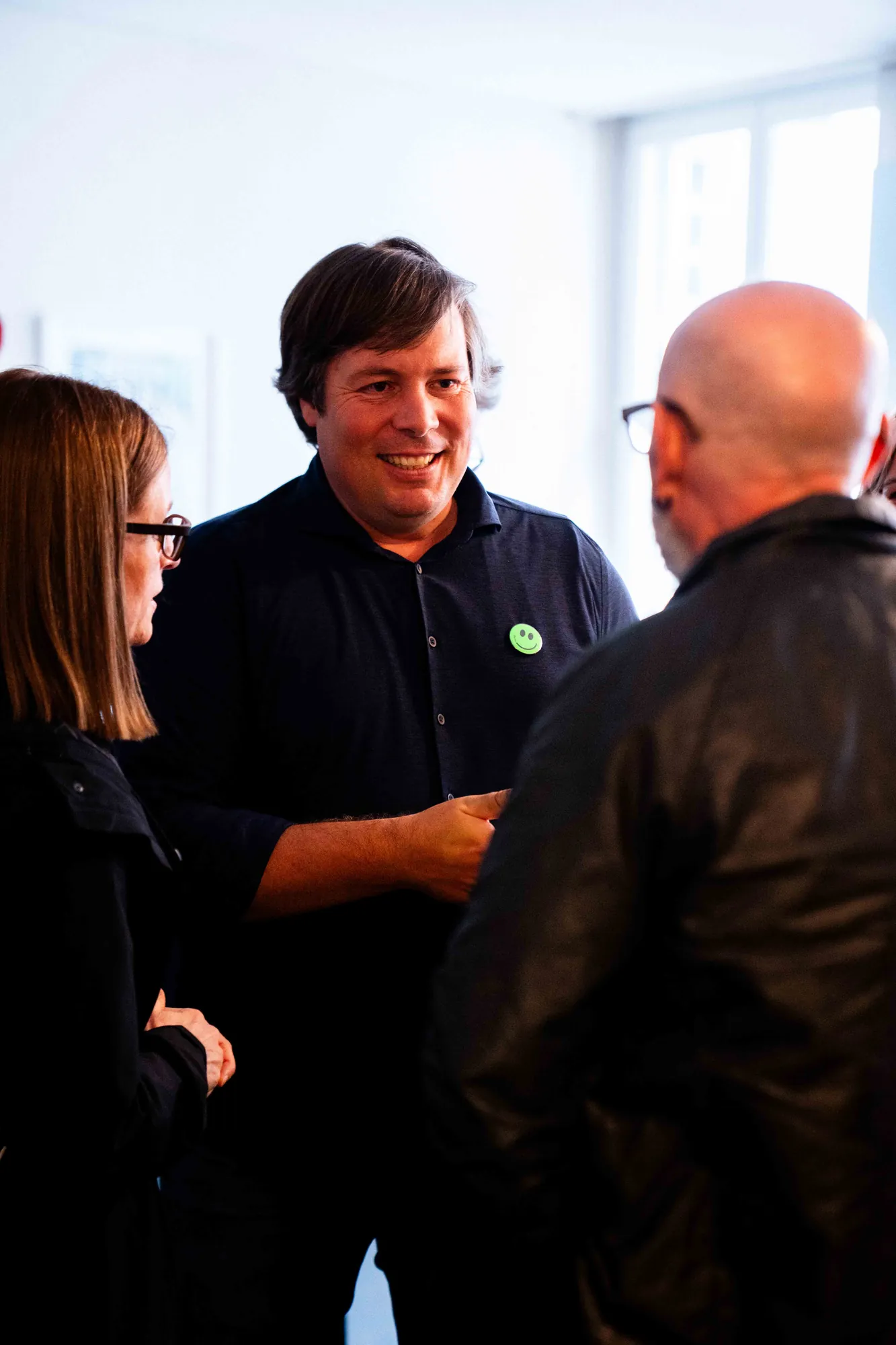 Gast im dunkelblauen Poloshirt mit grünem Smiley-Button lacht im Gespräch mit zwei Personen in einem hellen Raum.